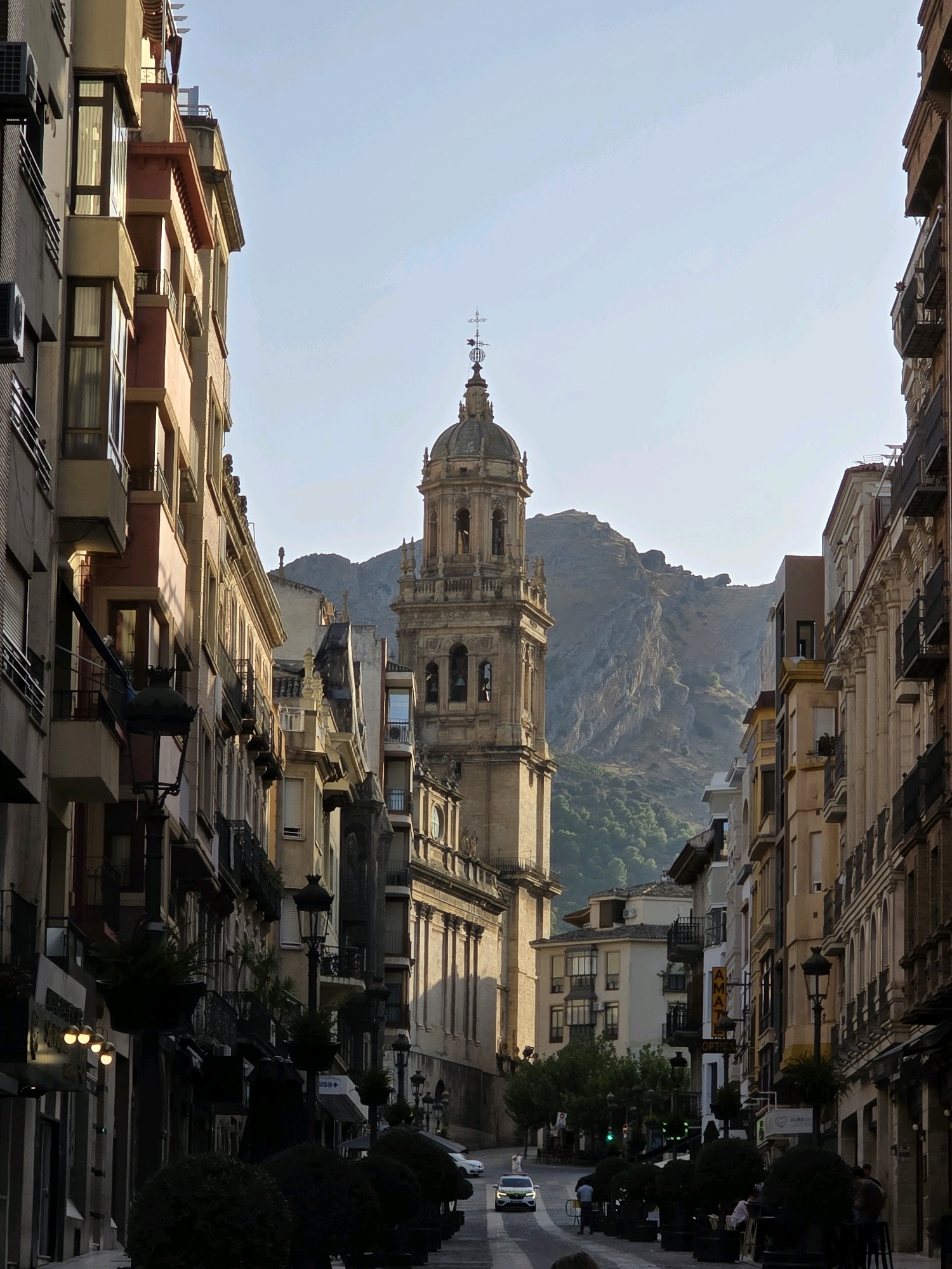 The image captures a street scene in Jaén, Spain, dominated by the Jaén Cathedral's bell tower, a prominent landmark. The tower, constructed of stone, features arched windows and a cross at its pinnacle. It is flanked by buildings of varying architectural styles, including a historical stone facade on the left and a multi-story building with balconies on the right. A steep, rocky mountain covered in sparse vegetation rises in the background, bathed in soft, late afternoon sunlight.
The street itself is paved and lined with shops and restaurants, indicated by signage and awnings. In the foreground, the street is partially obscured by the shadows of buildings and decorative potted plants. A white car is visible driving down the street, suggesting active use. A few individuals are discernible on the sidewalks, with one person standing near the car and others seated at outdoor tables, implying a relaxed atmosphere. The sky above is clear and pale blue, with a slight haze visible near the mountains. The overall impression is of a charming European city street during daylight hours.