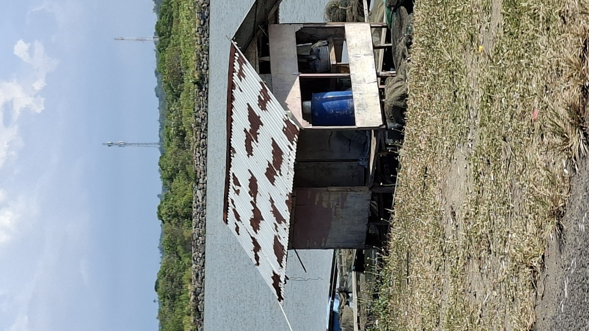 The image depicts a small, weathered shack situated on a body of water. The shack has a corrugated metal roof with visible rust spots and appears to be constructed from salvaged materials. Inside the open structure, a large blue barrel is prominent, along with some stored items. The shack is supported by wooden stilts and rests on a platform that is partially submerged or floating. The surrounding environment includes water, a rocky embankment, and lush green vegetation further inland. Two tall communication towers are visible in the distance, silhouetted against a cloudy sky. The foreground shows dry, sparse vegetation and a paved surface, possibly a road or pathway. The scene suggests a humble, possibly isolated dwelling by the water's edge. This appears to be located in Lhokseumawe, Indonesia, given the characteristic communication towers common in the area and the coastal environment. There are no people visible in the image.