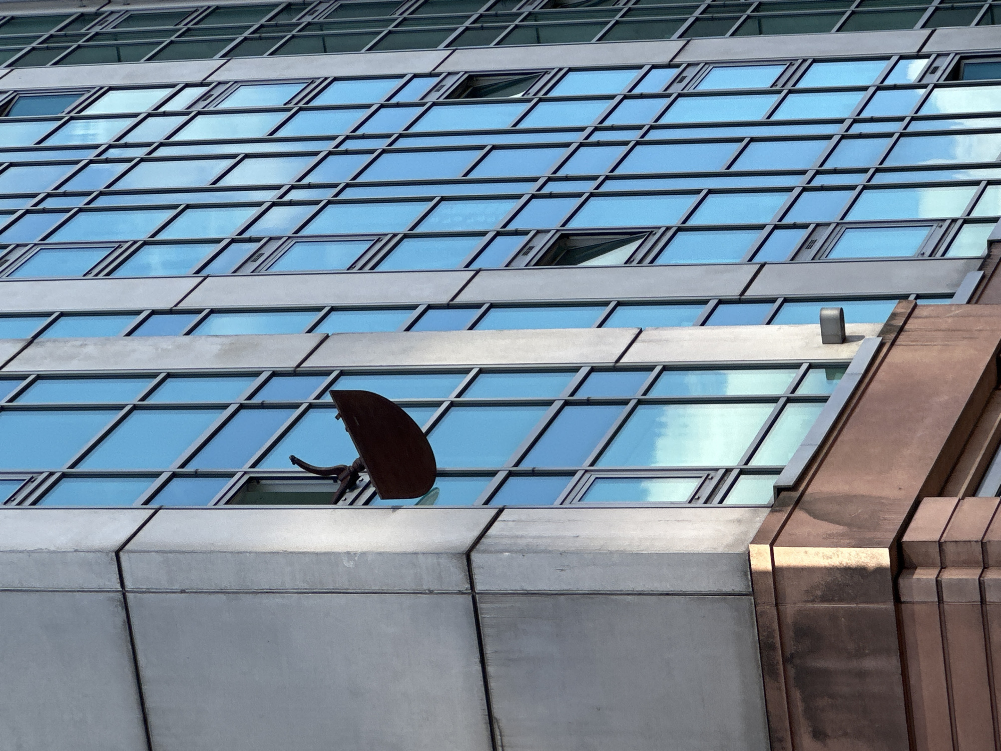 The image provides an upward, close-up view of a modern building's facade, set against a clear blue sky, likely in an urban environment such as London. The building is characterized by a repeating grid of rectangular glass panels that reflect the bright sky and scattered white clouds, indicating clear daytime weather. Several of these windows, particularly in the upper and mid-sections, are partially open, tilting outwards from their bottom edge.
Below the extensive glass sections, there are horizontal bands of light grey, matte concrete or metallic cladding. From one of the open windows on a lower section of the glass facade, a distinctive, dark, and irregularly shaped object protrudes. This object appears to be a sculpture or a unique architectural fixture, crafted from rusted or dark-colored metal, possibly cast iron or weathered steel. It features a broad, curved, and somewhat abstract main body, from which a more slender, organic, and arm-like extension reaches back into the open window.
To the right, a prominent vertical architectural element of the building is visible, constructed from a bronze or copper-toned material with a multi-faceted, stepped design, reflecting light differently across its surfaces. No people are present, and no text is visible anywhere in the image. The scene captures a blend of modern architectural linearity with an intriguing, contrasting artistic or structural element.