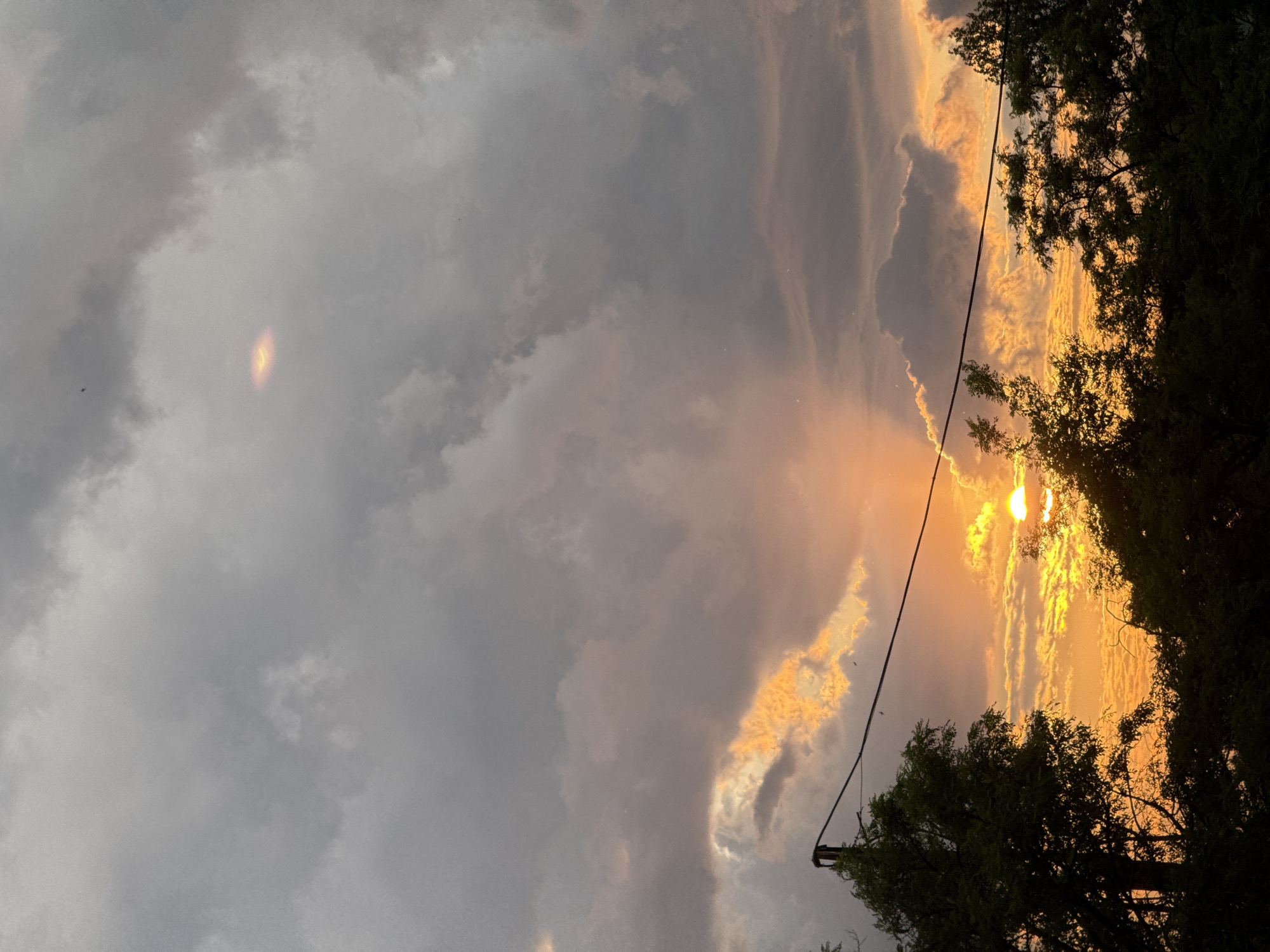 A sunset is depicted over a silhouette of trees, with dark clouds overhead tinged with orange and yellow from the setting sun. A utility pole with wires is visible on the left side of the frame. The sky is dramatic, with shafts of light breaking through the clouds. No people are present in the image.