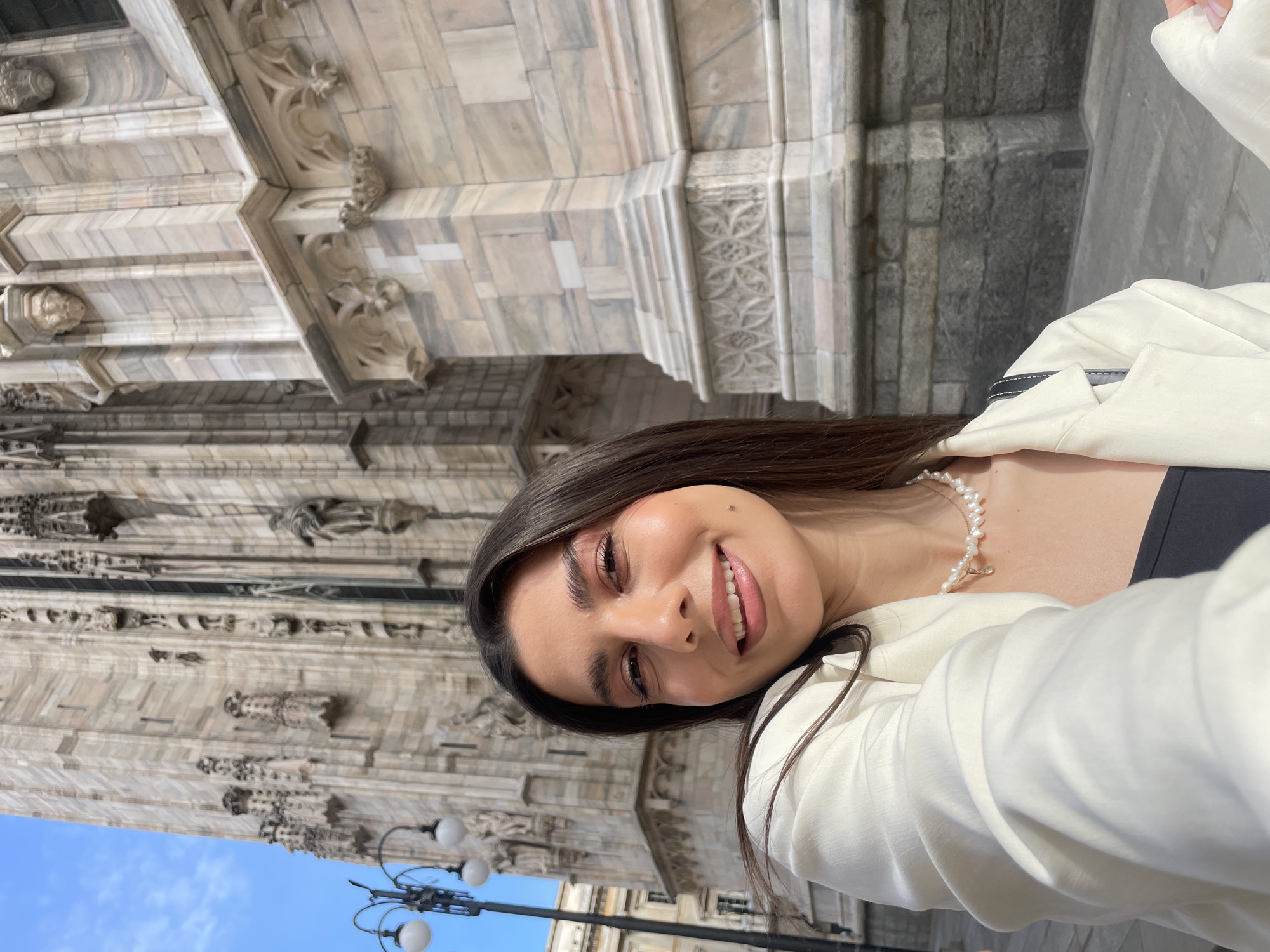 A young woman is posing for a selfie in front of the Duomo di Milano, a large cathedral in Milan, Italy. The cathedral's facade is visible behind her, with intricate stone carvings and architectural details. The sky is partially visible in the bottom left corner. The woman is wearing a white blazer over a dark top and a pearl necklace. She is smiling at the camera.