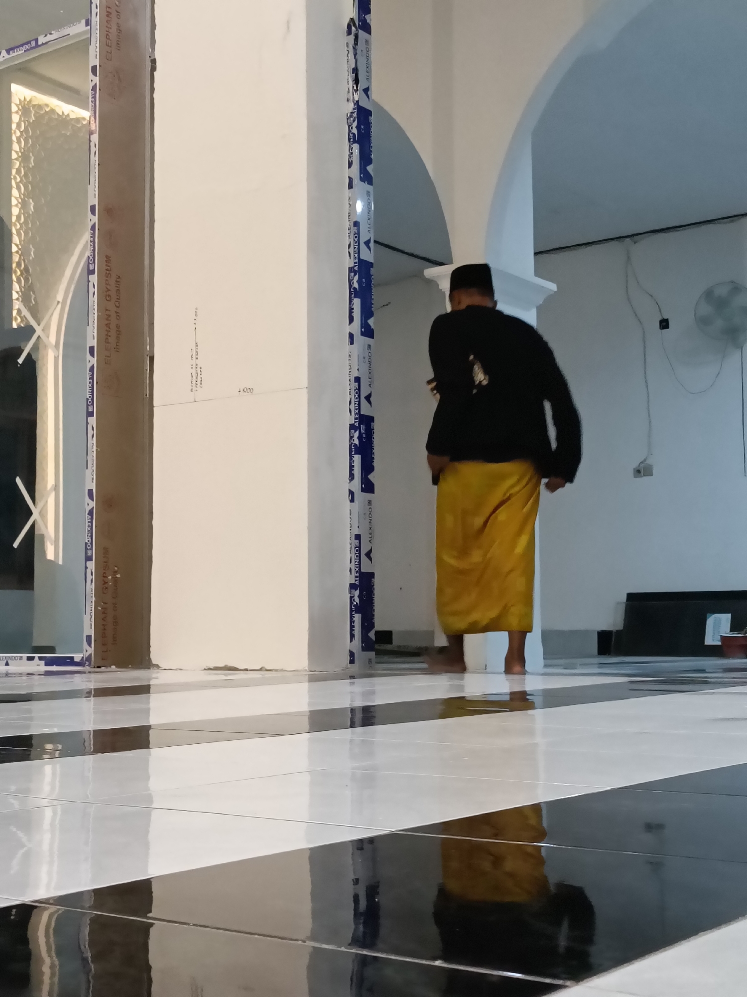 A person wearing a black long-sleeved shirt and a yellow sarong is walking across a black and white tiled floor. The person is facing away from the camera, towards the right side of the image. They are barefoot. The floor is highly reflective. In the background, there are white columns and arches. There is a fan on the wall in the upper right corner of the image. The scene appears to be indoors, possibly a mosque or a public building. The provided crops confirm details of the person's attire and the architectural elements. The scene does not contain any specific landmarks that would allow for precise location identification within Kediri City, Indonesia, beyond it being an indoor setting in that region.