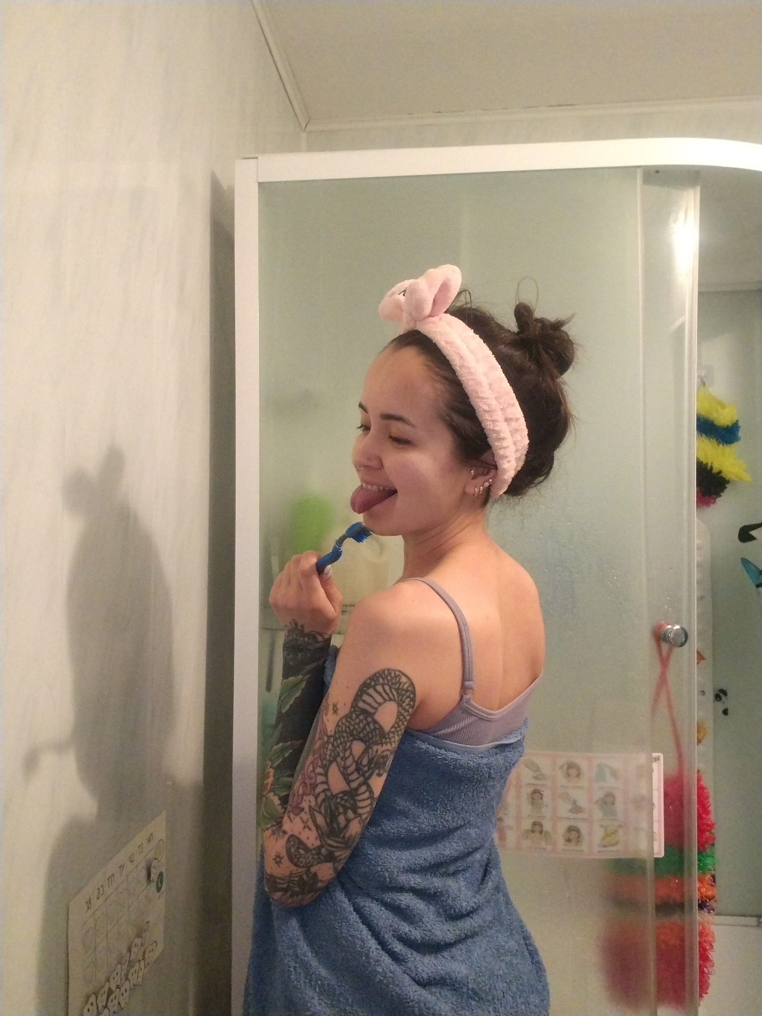 A woman with fair skin stands in a bathroom, partially covered by a blue towel. She is wearing a light purple sports bra with thin straps and a pink headband with bunny ears. Her dark hair is styled in a messy bun. She has multiple piercings in her left ear and a large tattoo covering her left arm. The woman sticks her tongue out, holding a blue toothbrush to her mouth.
The bathroom features a shower with a frosted glass door. A calendar with cartoon figures is attached to the shower door. A wall calendar with small skull-shaped magnets is affixed to the wall next to the shower. The wall is pale, and the room is adequately lit.
There is no indication that the photograph was taken in the city of Noviy, Russia.