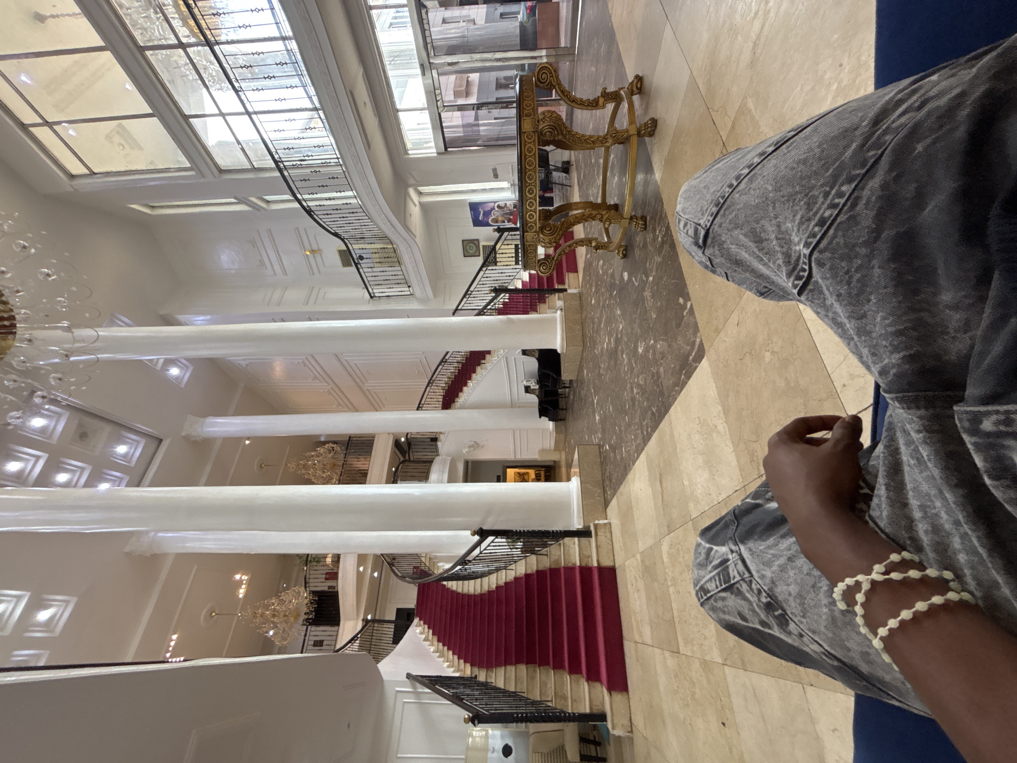 The image captures a wide-angle view of a grand hotel lobby in Owerri, Nigeria. A person's legs and arm, adorned with a white beaded bracelet, are in the foreground, suggesting the viewer is seated on a blue couch. The lobby features a prominent, sweeping red carpeted staircase that curves upwards, flanked by large white columns. Ornate chandeliers hang from the high ceiling, and elegant railings line the upper balconies and staircases. A decorative golden table with intricate carvings sits in the middle ground, near large glass doors that lead outside. The floor is tiled in a combination of beige and brown marble. The overall impression is one of opulence and spaciousness.
