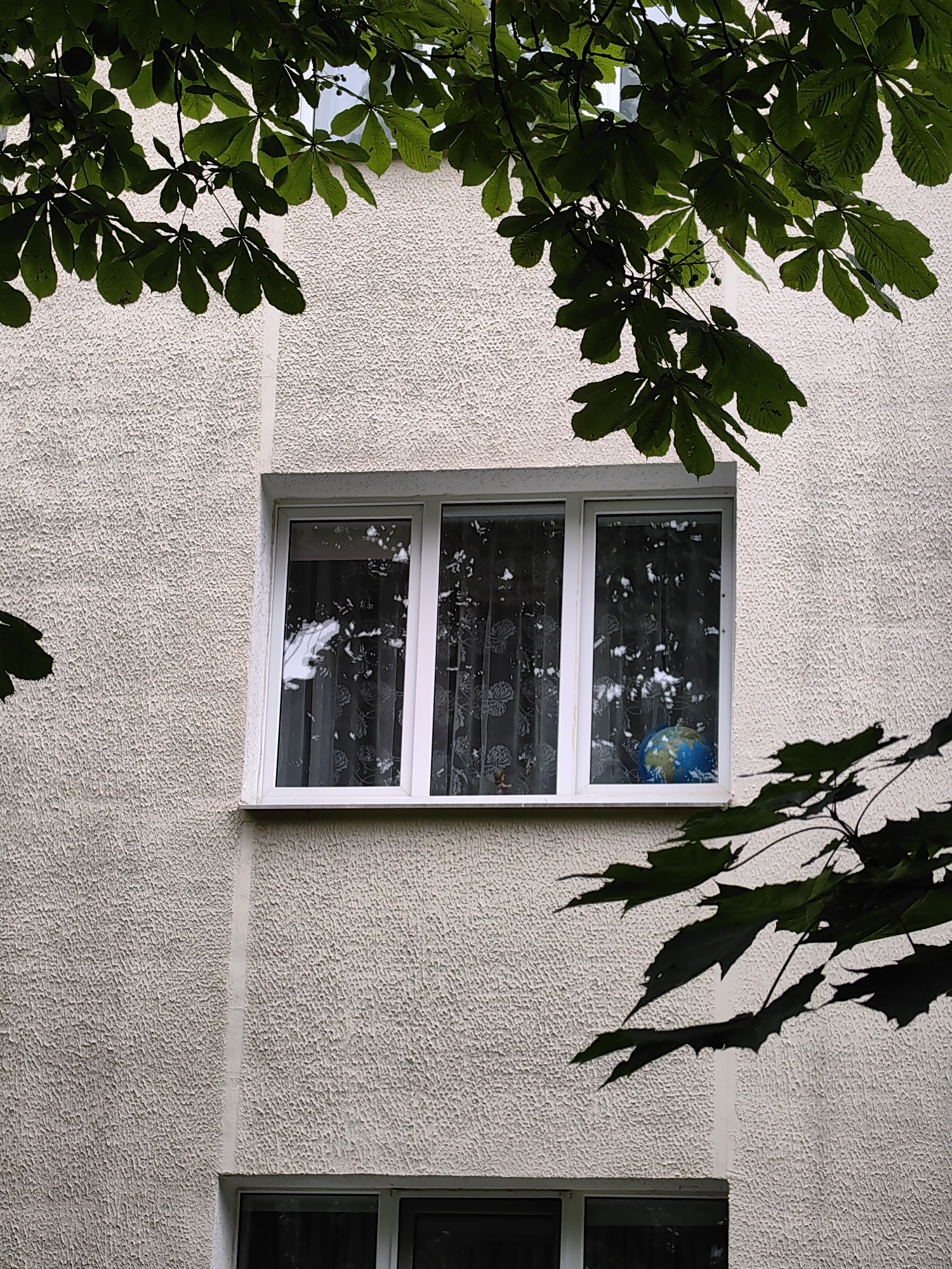 A medium shot captures the exterior of a building in Minsk, Belarus, with green leaves from a tree casting shadows on the textured beige wall. A white-framed, triple-pane window is centrally located, revealing sheer white curtains with a floral pattern. Inside the rightmost pane, a small blue and green globe is visible. The lower portion of the image shows the top of another window.