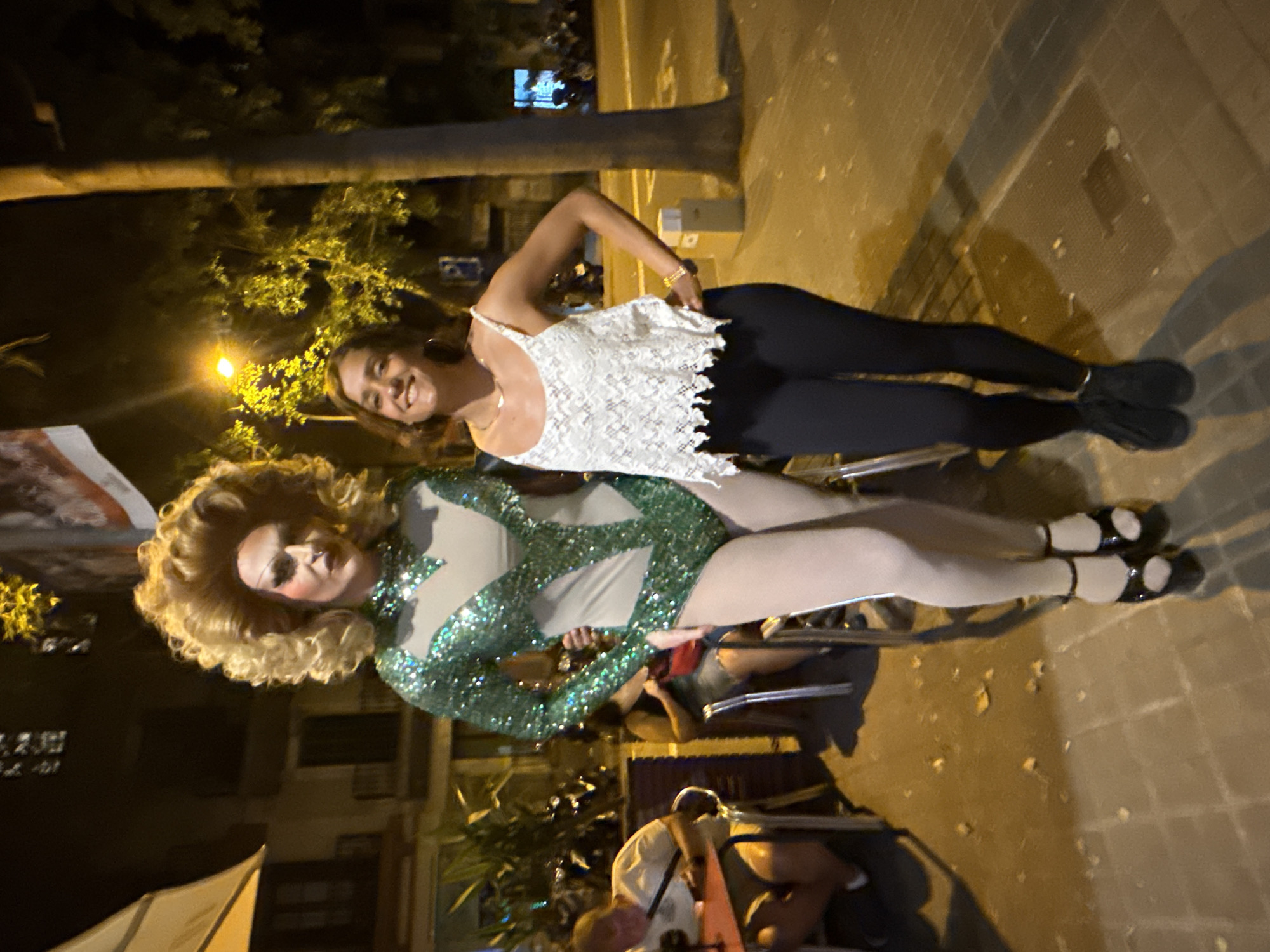 In Barcelona, Spain, two individuals pose for a photograph at night on a sidewalk next to an outdoor seating area for a restaurant or bar. On the left, a person in drag wears a sparkling green sequined costume with a cutout design, white sheer hose, and black heeled sandals. They have a voluminous blonde wig, heavy makeup including false eyelashes and a nose ring. To the right, a smiling woman in a white lace top and black leggings stands with her hand on her hip, wearing a gold necklace and bracelet. Behind them, trees are visible, and streetlights illuminate the scene, casting shadows on the tiled pavement. In the background, patrons are seated at tables, and street activity can be seen, including parked motorcycles.