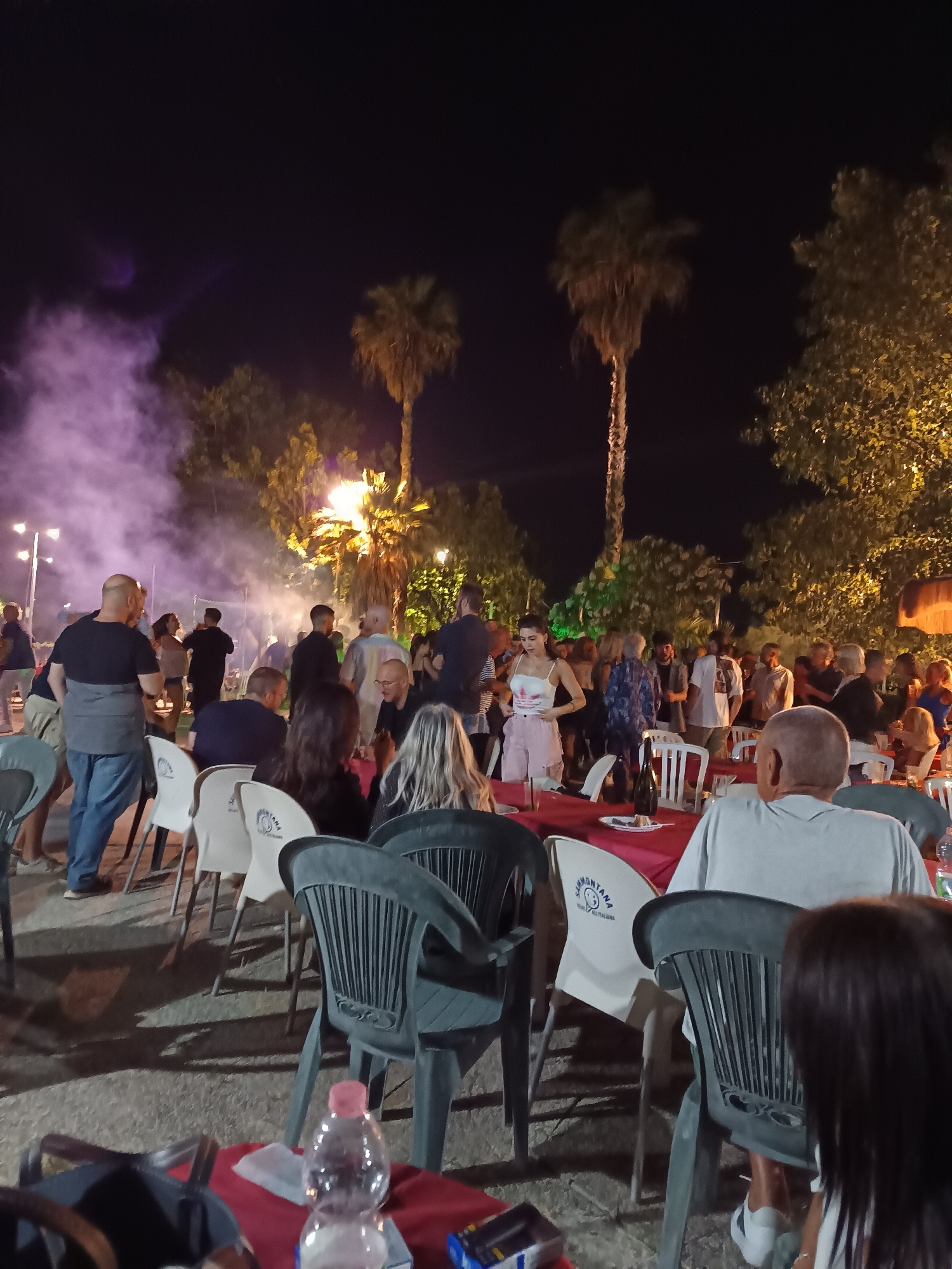 This image depicts an outdoor gathering at night in Giulianova, Italy. The scene appears to be a communal dining or social event, possibly a festival or celebration, taking place in a paved outdoor area with trees and palm trees. In the foreground, several tables covered with red tablecloths are set with chairs, some of which are occupied by people dining or socializing. A bottle of water is visible on one of the tables. In the background, a crowd of people are mingling, some dancing. There is a significant amount of smoke or mist illuminated by lights, creating a dramatic atmosphere. The overall ambiance suggests a lively and social evening event.