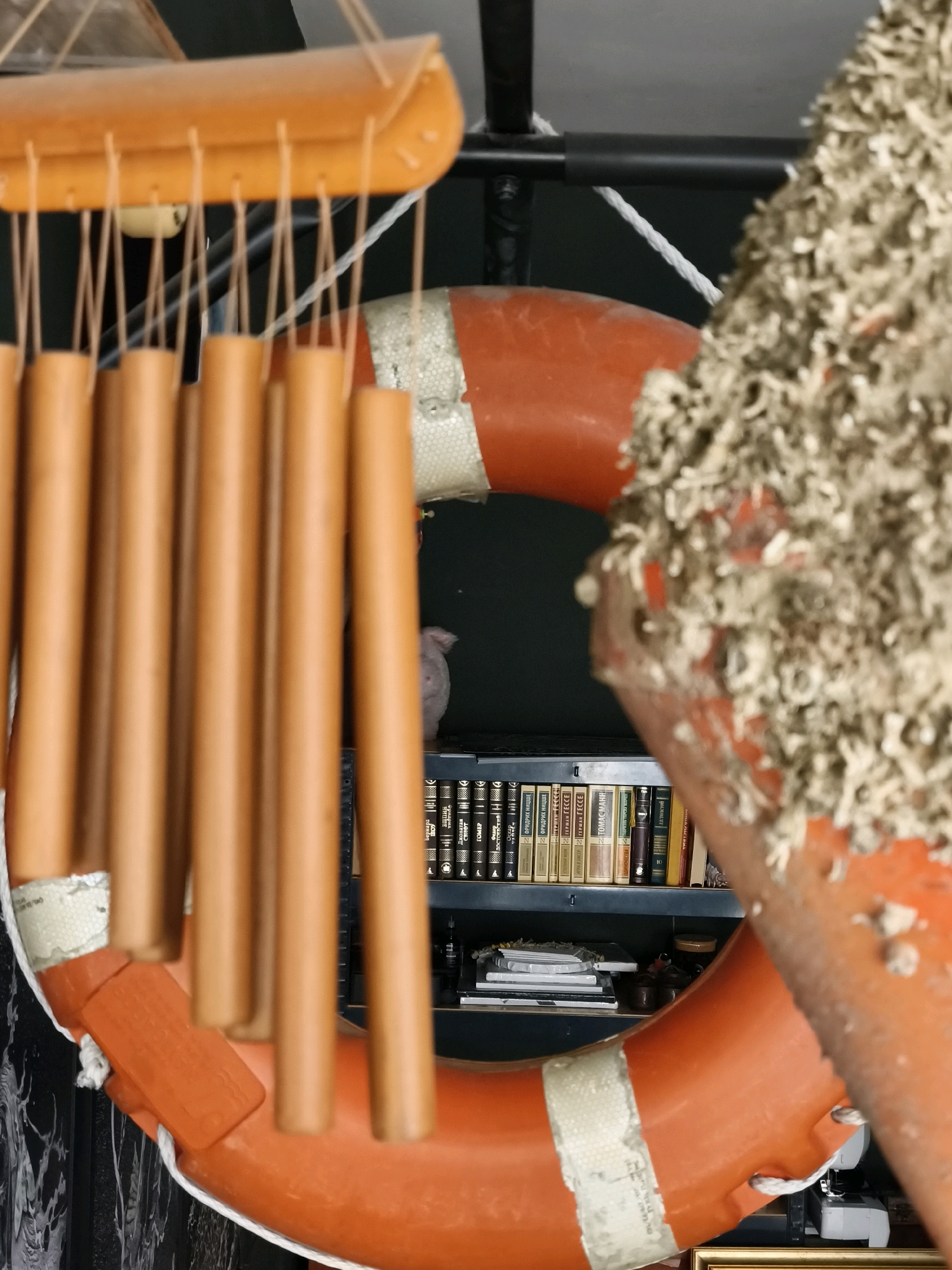 The image shows a close-up of decorative elements, likely in an outdoor setting, potentially related to a nautical theme. In the foreground, several light brown, cylindrical wind chimes hang from a light brown wooden frame. Behind the wind chimes, a bright orange life preserver is prominently displayed, partially covered in what appears to be dried plant matter or debris. A white rope is visible tied around the life preserver. Further in the background, a shelf unit is visible, filled with books and other assorted items. The overall impression is one of a weathered or bohemian aesthetic, possibly in a coastal area.