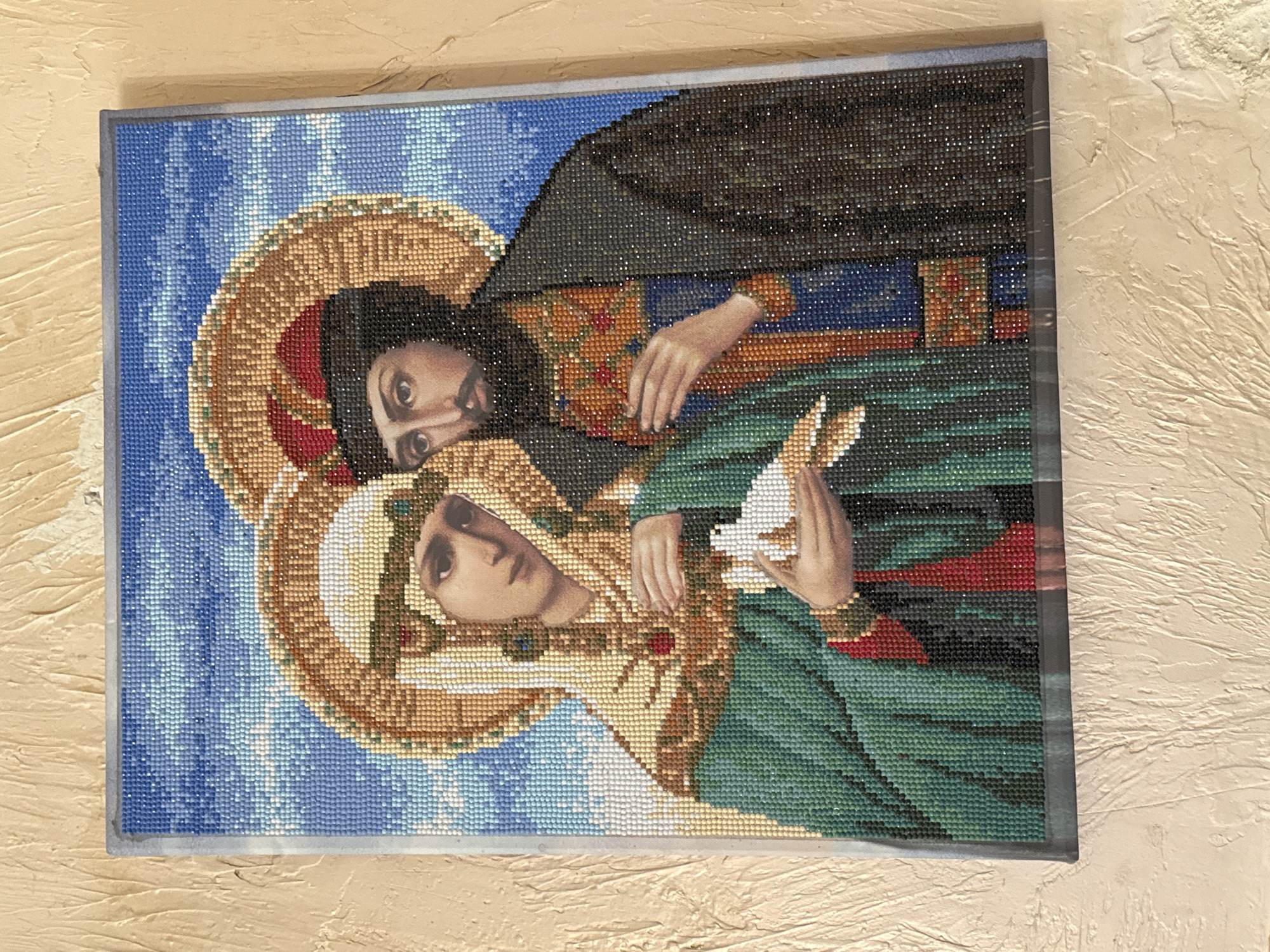 An intricate, framed artwork, likely a diamond or bead painting, depicts two religious figures against a mosaic blue background. The artwork is photographed on a textured, light beige wall, and is rotated 90 degrees clockwise from its natural upright orientation.
The primary subject is a female figure positioned in the foreground. She wears a green garment and a white head covering, and holds a small white bird, possibly a dove, in her hands which are clasped near her chest. Her face, realistically rendered, looks subtly downwards and to the left, emanating a serene expression. A prominent, ornate golden halo encircles her head.
Behind her, partially visible and looking over her right shoulder, is a male figure. He has dark hair, a beard, and wears a dark cloak over a richly decorated garment of red, blue, and gold tones. His face, also realistically rendered, gazes directly forward with an intense expression. He too has an elaborate golden halo.
The unique aspect of the artwork is the contrast between the smoothly painted or printed faces and hands of the figures, and the rest of the image, which is composed of countless small, glittering beads or diamond facets. This technique gives the garments, halos, and the blue background a pixelated, shimmering effect, suggesting a starry sky or divine radiance. No text is visible in the image.