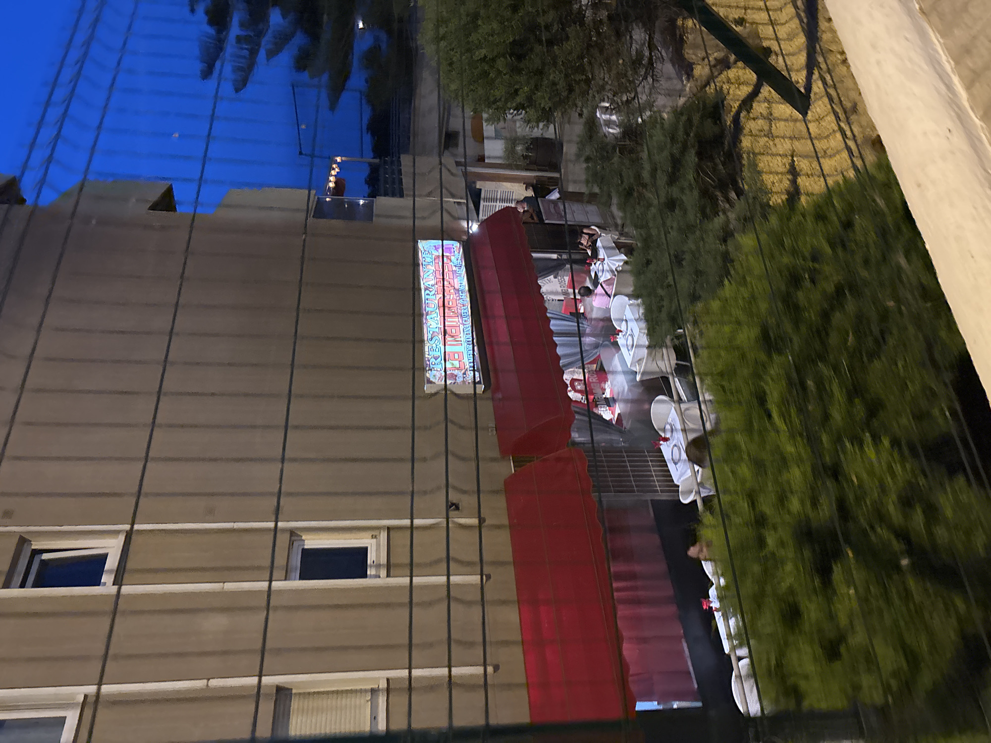 In Calp, Spain, a restaurant with red awnings is visible at dusk. The restaurant's name, "RESTAURANTE LE INDIENNE," is displayed on a neon sign. Several tables with white tablecloths are set outside, some with diners. The building appears to be an apartment complex, with windows visible on the upper floors. A fence runs across the foreground, partially obscuring the view. Lush green foliage is present in the lower portion of the image. The sky is a deep blue, indicating evening.