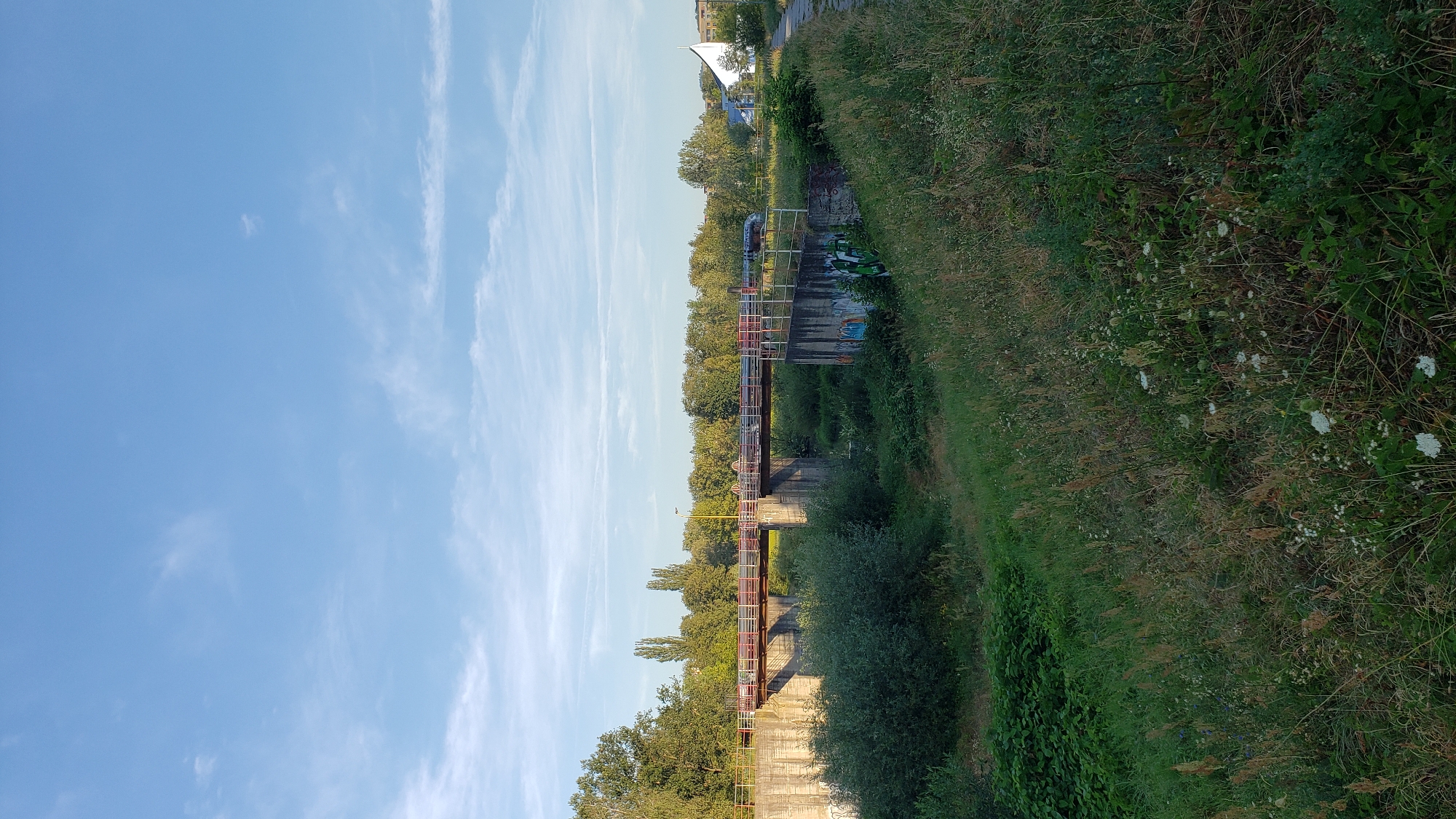This media file depicts a scene of a bridge over a waterway, with lush greenery and a blue sky. The bridge has a reddish-brown railing and appears to be made of concrete and metal. It crosses what seems to be a canal or a small river.
On the left side of the image, a vast expanse of blue sky is visible, with thin, wispy clouds. The sky suggests it is daytime, likely late afternoon or early evening given the warm lighting on the vegetation.
On the right side, a dense embankment covered in green grass and wildflowers slopes upwards. The vegetation is tall and appears somewhat wild, indicating a natural or semi-natural setting.
In the background, beyond the bridge and the trees lining the waterway, there are some buildings and a distinctive white, tent-like structure, possibly part of a modern building or a park feature. There are also some signs of human presence, such as graffiti on the bridge supports and some piping running along the bridge.
The overall impression is one of a tranquil outdoor scene, likely a park or a recreational area in Prešov, Slovakia. There are no people visible in the image, and the scene evokes a sense of peace and natural beauty. The time of day appears to be sunny, with a clear sky and soft light.