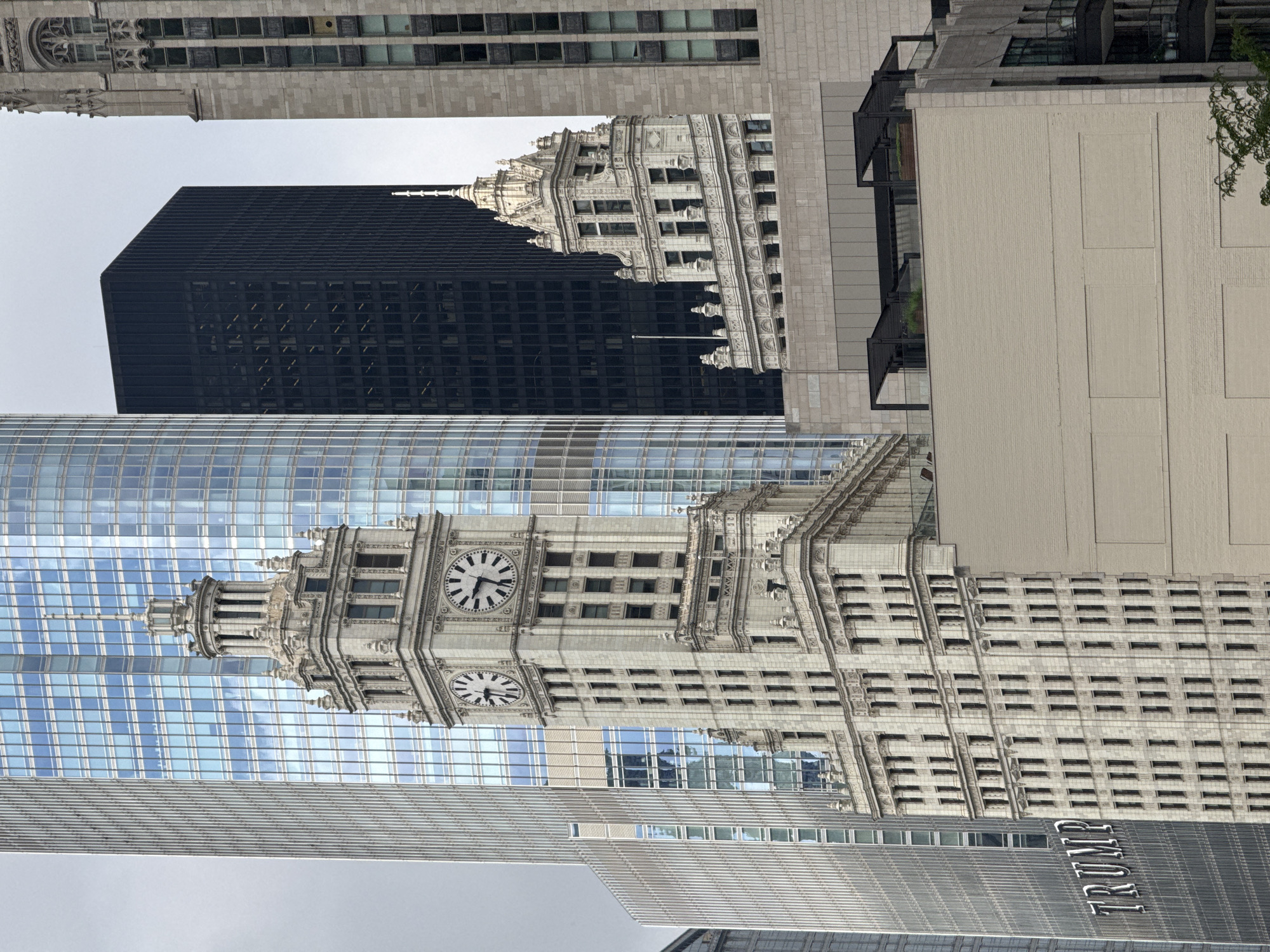 The photograph captures a close-up view of several skyscrapers in Chicago, Illinois, United States of America. The most prominent structure in the foreground is the historic Wrigley Building, characterized by its ornate white stone facade and twin clock towers. To its right, a section of a more modern, grey stone skyscraper is visible. Behind the Wrigley Building, a tall, glass-fronted skyscraper with a blueish tint dominates the background. To the left of the Wrigley Building, a sleek, modern skyscraper with a metallic facade, bearing the "TRUMP" logo, is partially visible. Further in the background, a dark, rectangular skyscraper stands out, contrasting with the lighter tones of the other buildings. The sky is overcast, casting a muted light over the urban landscape. The image is a detailed snapshot of the city's diverse architectural styles, showcasing a mix of historical and contemporary high-rise buildings.