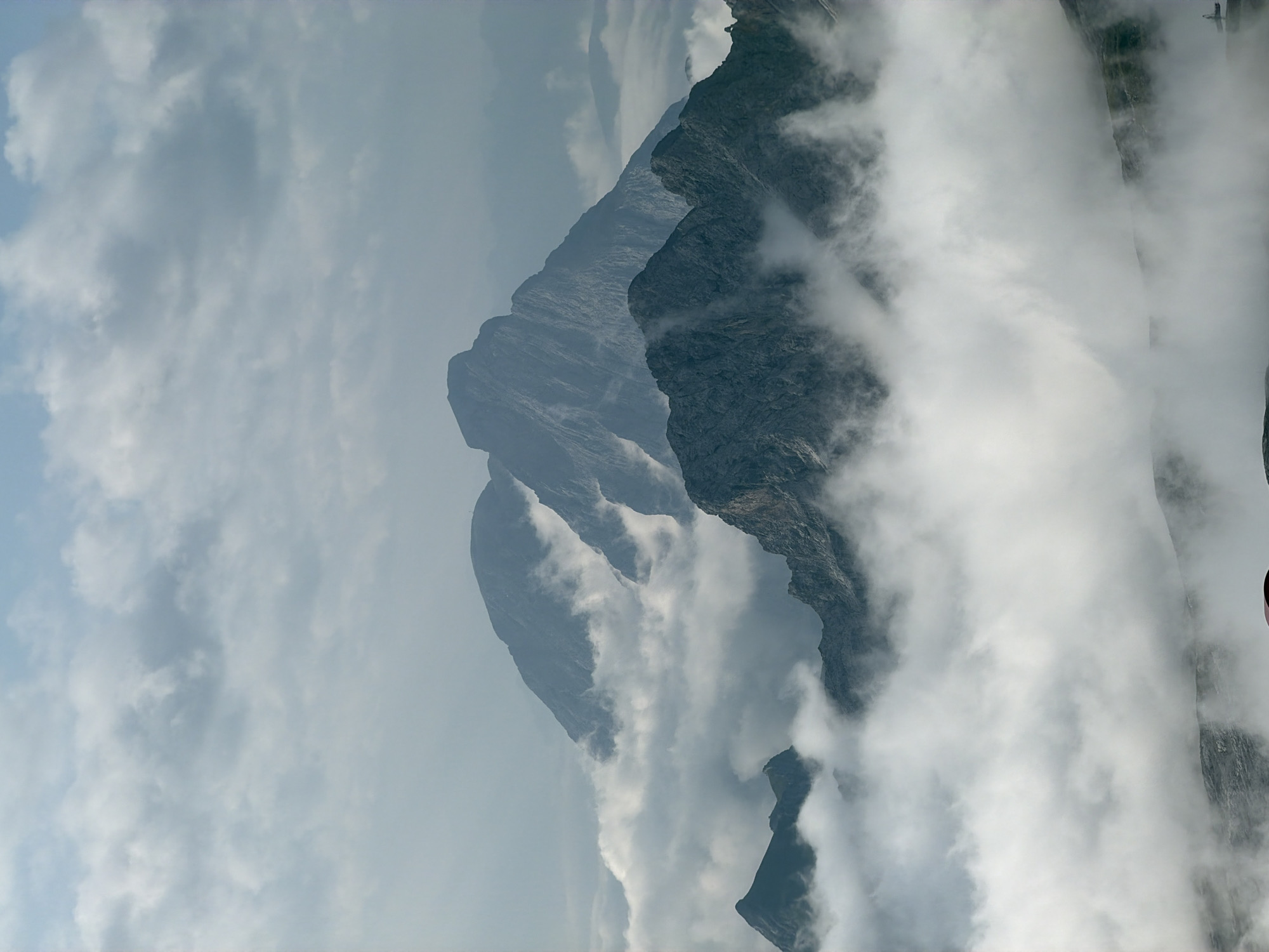 This image depicts a mountainous landscape partially obscured by clouds. The prominent features are several rugged mountain peaks, rendered in shades of grey and blue. The mountains are situated at varying distances, creating a sense of depth. A thick layer of white, wispy clouds blankets the lower portions of the mountains, giving the impression that they are emerging from or sinking into a sea of mist. The sky above is a pale blue, with lighter, scattered clouds. The overall atmosphere is serene and somewhat dramatic due to the interplay of light and shadow on the mountains and the pervasive cloud cover. While the image captures a natural environment, it does not provide any specific visual cues to pinpoint a location within Garmisch-Partenkirchen, Germany, nor does it contain any discernible people, objects, or actions.