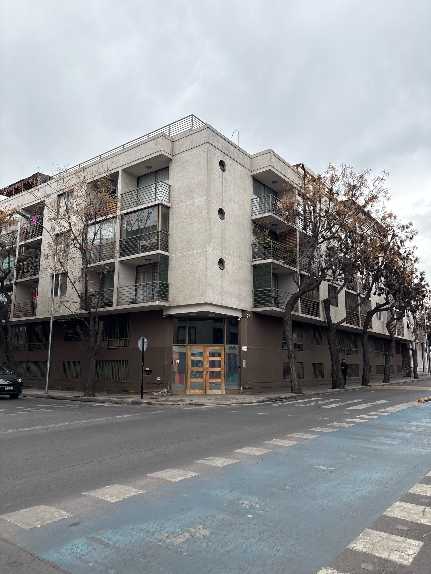 An exterior, medium-angle, eye-level, vertical shot shows a modern, multi-story apartment building on a street corner in Santiago, Chile. The building has a light-colored, textured facade with several balconies, large windows, and some circular decorative elements. Bare trees with brown foliage are planted along the sidewalk in front of the building, obscuring some of the lower floors. The street in the foreground has a pedestrian crosswalk with blue and white painted markings. A dark-colored car is parked on the left side of the street, and a person in dark clothing stands on the sidewalk on the right side of the street, facing the building. The sky is overcast and gray.