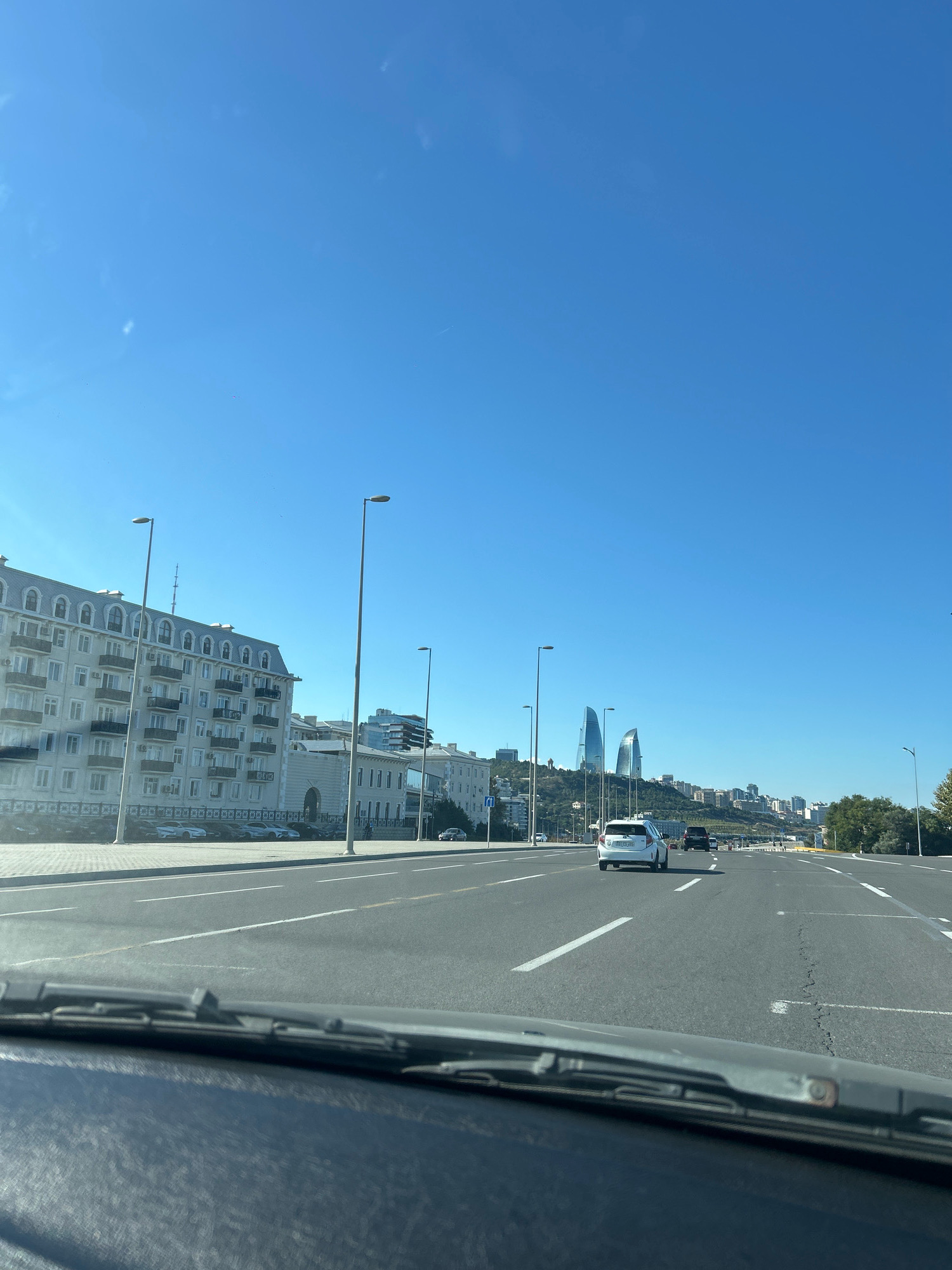 A street view from a vehicle traveling on a multi-lane road, looking ahead towards a cityscape. The sky is clear blue. On the left side of the road, a light-colored, multi-story building with a mansard roof and balconies is visible. Further down the street, other buildings and a hill with modern skyscrapers, including two flame-shaped towers, are prominent. Several cars are on the road, some moving and some parked. Streetlights are spaced along the road. The foreground shows the dashboard and windshield wipers of the vehicle. The scene appears to be in Sabail Raion, Azerbaijan, as evidenced by the architecture and the distinct flame-shaped towers of the Flame Towers, a landmark of Baku.