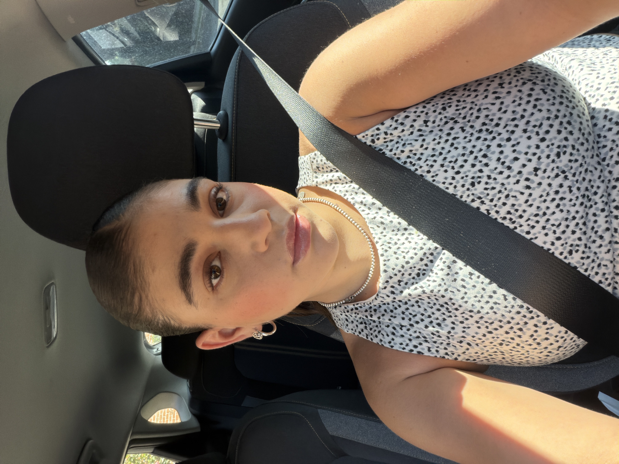 A woman is in a car wearing a seatbelt. She is wearing a patterned shirt and a silver necklace. She is looking at the camera. The location is implied to be in or near San Bernardino Tlaxcalancingo, Mexico.