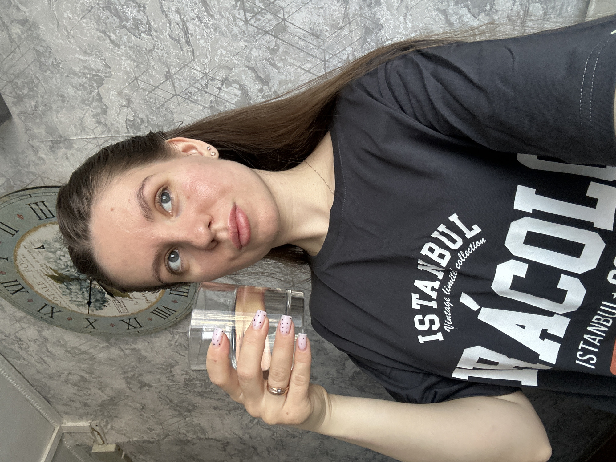 A woman is holding a glass in her hand. She is wearing a black t-shirt with the word "ISTANBUL" printed on it. She has long, brown hair and her nails are painted with a light pink base with black polka dots. She is wearing a ring on her ring finger. Behind her, there is a wall with a patterned gray wallpaper. To the left of the woman, there is a clock. The exact location in the city of Klin, Russia is not discernible.