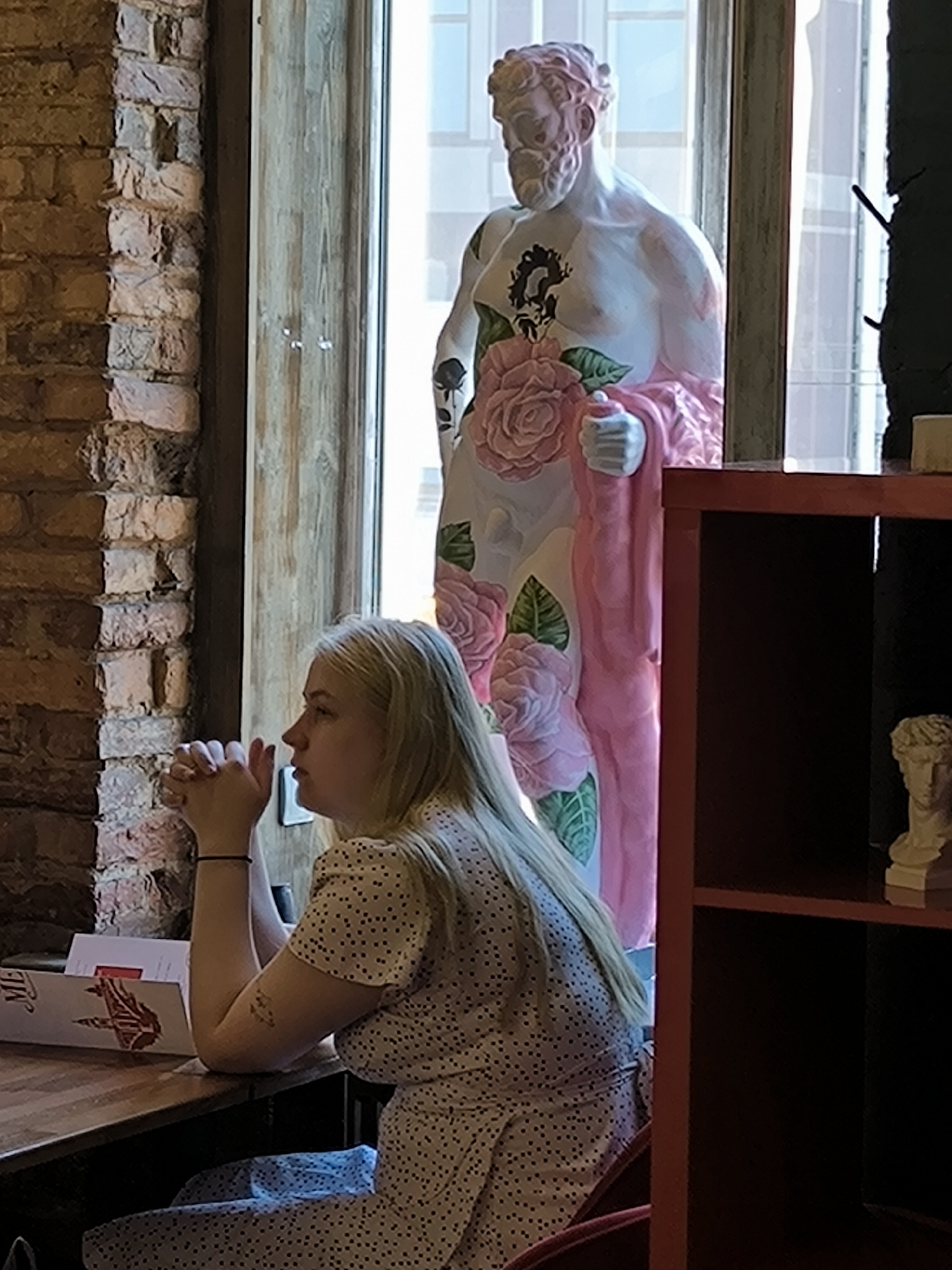 The image depicts an indoor scene within a cafe or restaurant, identified as being in Saint Petersburg, Russia, due to an illustration of Saint Isaac's Cathedral on a menu partially visible on a wooden table. A young woman with long blonde hair is seated in profile on the left side of the frame, looking towards the right. Her hands are clasped on the table, and she wears a light-colored dress with small dark polka dots. A thin black bracelet is present on her left wrist, and a small, indistinct tattoo is visible on her right upper arm. To her left is an exposed brick wall. Behind her, separated by a light-colored wooden window frame, a large white male classical-style statue is visible. The statue features painted pink roses and green leaves on its torso and legs, a dark, abstract design on its chest, and a pink drape over its lower body and left arm. Beyond the statue, through the window, faint building outlines are visible under natural light. On the right side of the frame, a reddish-brown wooden shelving unit with a dark interior is partially visible, displaying a small white classical bust on its top shelf.