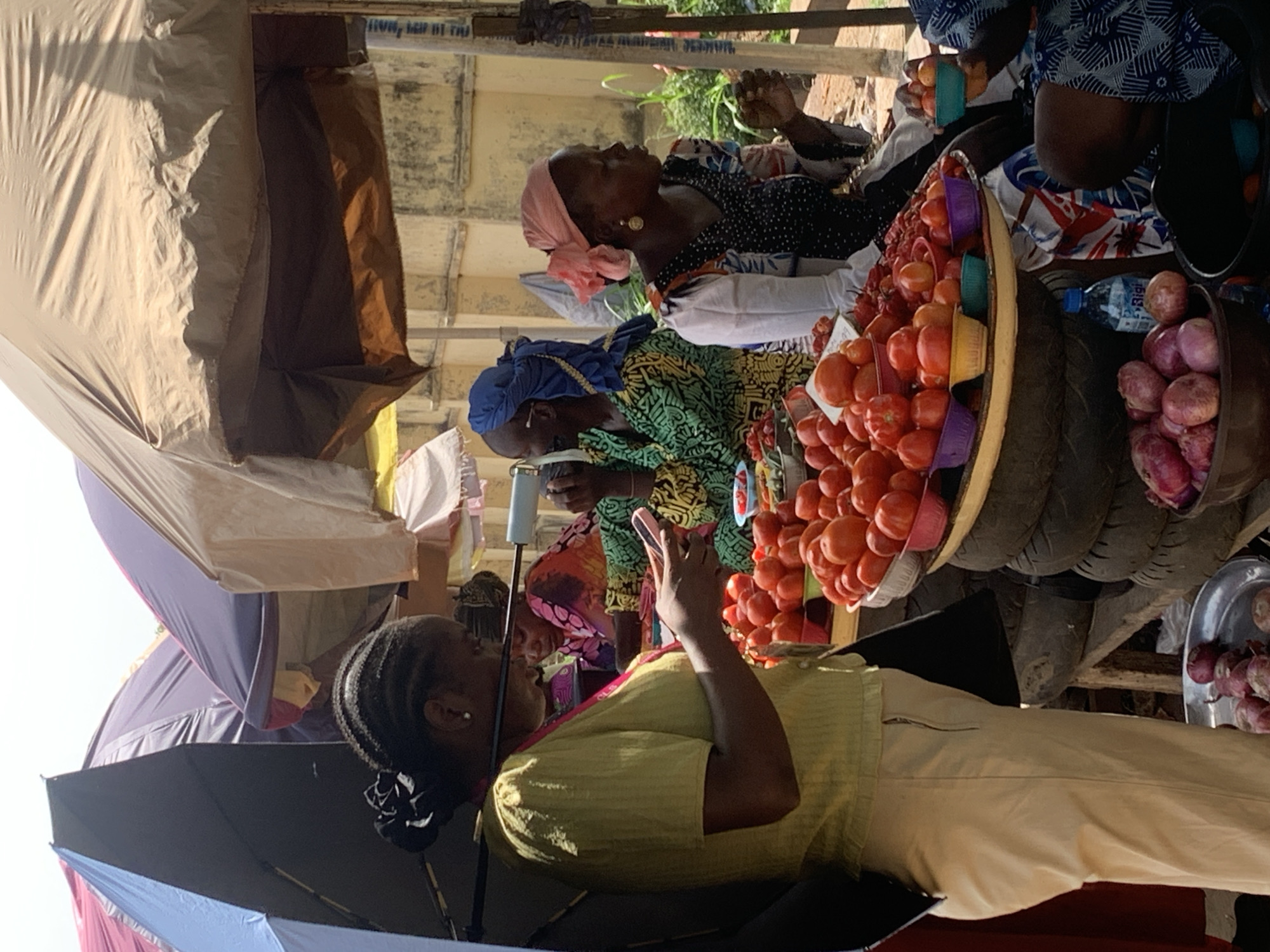 This image depicts a bustling outdoor market scene in Ilorin, Nigeria, under bright daylight. Several women are positioned within their stalls, which are covered by large tarpaulins and open umbrellas to provide shade from the sun.
In the foreground, a woman wearing a light yellow top and cream-colored pants is seated, looking down at a smartphone held in her hands. Her braided hair is styled neatly, and she is partially sheltered by a black umbrella. Behind her, another woman, dressed in a vibrant green and black patterned shirt and a blue headwrap, is also seated and appears to be looking down, possibly at a phone or her merchandise.
A prominent feature in the mid-ground is a large, circular, flat basket overflowing with numerous ripe red tomatoes. Small, colorful plastic bowls (purple, green, blue) are nestled among the tomatoes. This display rests upon a stack of dark-colored tires, serving as an improvised stand. Next to the tomatoes, a container holds a pile of red onions, and a blue plastic bottle of "Bigi" brand drink is visible.
Further back, to the right, a third woman is seated, wearing a black and white patterned top and a light pink headwrap, accessorized with gold earrings. She is looking slightly upwards or to the side, with a calm demeanor. Portions of other market structures and people are faintly visible.
The background reveals a weathered concrete wall with faint, faded black text, including partial phrases like "HVA, MEP MI THJ" and "JATAWALE AMI...", suggesting old signage or graffiti. Some green foliage is also visible beyond the market structures. The scene conveys the daily life and commerce of a vibrant local market, with vendors tending to their goods and taking moments for themselves.