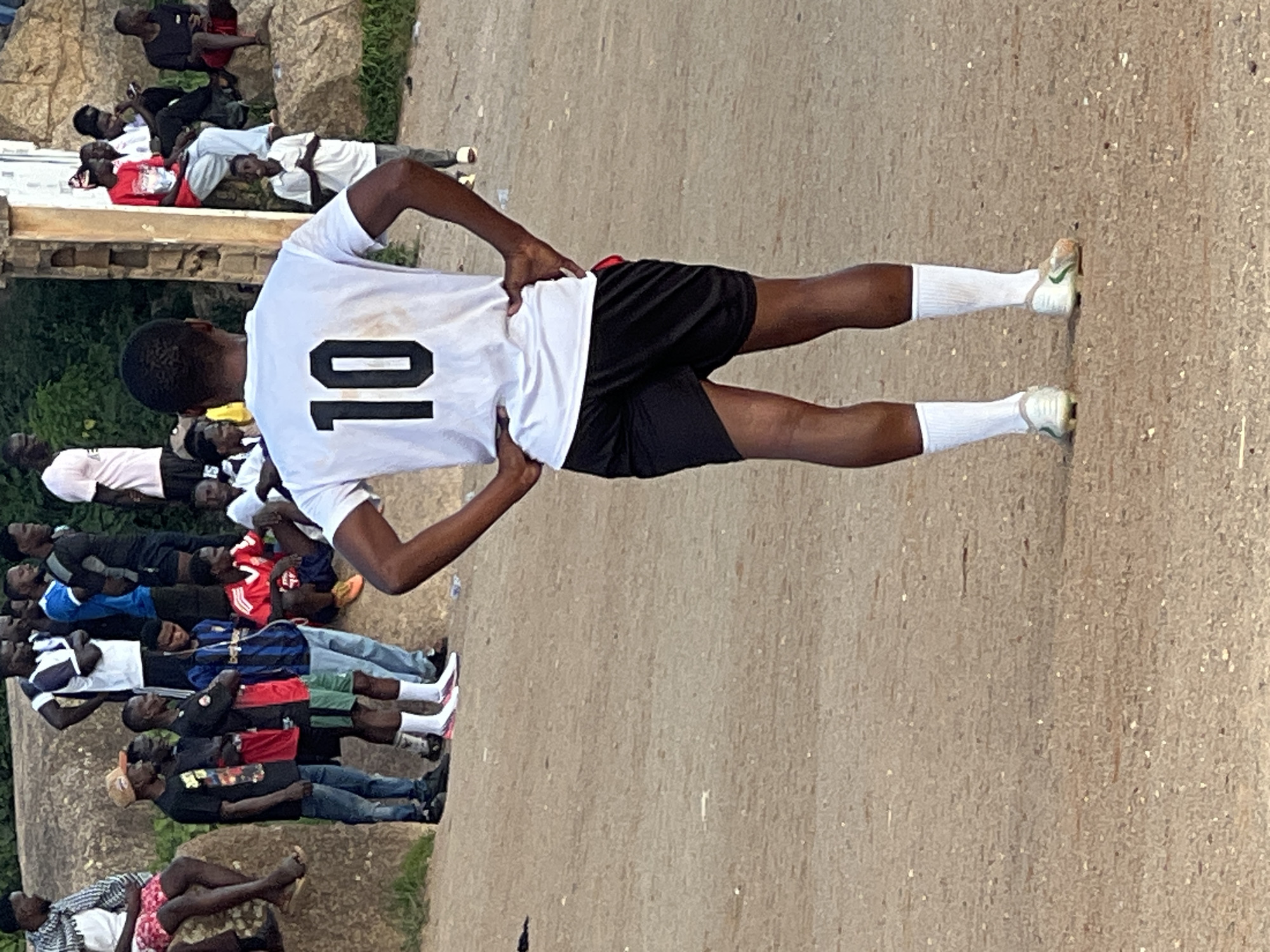 In the city of Jos, Nigeria, a young Black male soccer player stands facing away from the camera, hands on his hips. He is wearing a white t-shirt with the number "10" in black on the back, black soccer shorts, white socks, and soccer cleats. He is on a dirt field, likely a soccer pitch, with a crowd of spectators in the background. The spectators are also Black males, some seated on rocks and others standing. They are dressed in various casual clothing, including t-shirts, shorts, and jeans. The environment is outdoors, with some vegetation and stone structures visible in the background. The lighting suggests it is daytime, possibly late afternoon.