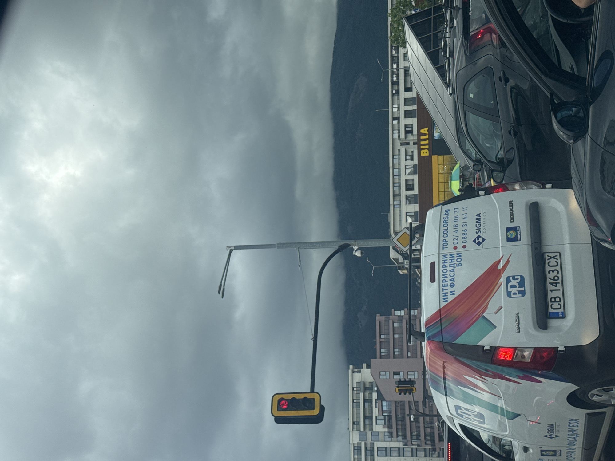 The image captures a daytime scene of urban traffic under an overcast sky, likely indicating a gloomy or potentially rainy day. The perspective is from inside a vehicle, looking forward into a traffic jam or cars stopped at a red light.
In the foreground, a white Dacia van with colorful graphics is prominently featured, displaying various brand names and contact information. Its rear license plate, "BG CB 1463 CX", confirms the location as Sofia, Bulgaria. Behind the Dacia van, a black car, possibly a Toyota based on roof racks, is also visible. Other vehicles are hinted at in the traffic.
To the right, a multi-story building with a yellow "BILLA" sign indicates a supermarket. Beyond the buildings, a large, dark mountain range dominates the background, partially obscured by heavy grey clouds that stretch across the sky. A traffic light, displaying a red signal, hangs from a pole, with other street infrastructure like traffic signs also visible.
Visible text on the white Dacia van includes "ИНТЕРИОРНИ И ФАСАДНИ БОИ" (Interior and Exterior Paints), "ТОР COLORS.bg", phone numbers "02/418 08 37" and "0886 31 44 17", and branding for "SIGMA COATINGS", "PRIMALEX", "PPG", and "DOKKER". The "BILLA" sign is clearly visible on the supermarket building.