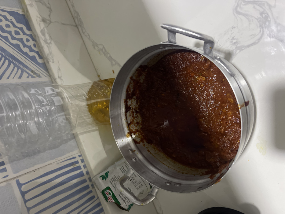 The image displays a close-up indoor scene, likely in a kitchen or food preparation area. The primary focus is a metallic pot, silver in color, tilted on its side, revealing a significant amount of dark reddish-brown, thick liquid or paste-like substance inside. This substance has a somewhat textured, lumpy appearance, consistent with a stew, curry, or sauce. The pot has two small, metallic handles on opposite sides.
To the left of the pot, a clear plastic bottle is visible. The bottle is partially filled with a golden yellow liquid, which appears to be cooking oil. The top part of the bottle seems to be cut or modified, suggesting it might be used for pouring or as a makeshift container. The bottle is lying horizontally on a surface.
Below the bottle and partially obscured by the pot, there is a small white packet with green text. Visible text on the packet includes "Dew", "Ingredients:", and "Sugar Free". Further down, the text "Ingredients: Veggies, Potato" can be partially deciphered, suggesting it might be a seasoning mix or an instant food packet.
The surfaces in the image consist of different materials. To the left, there's a countertop or wall with blue and white striped tiles arranged in a wavy pattern. Above this, and extending behind the pot, is a white surface with subtle grey marble-like patterns, possibly a countertop or backsplash. There are a few small splashes or stains of the reddish-brown substance from the pot on the white surface near the pot.
The lighting suggests an indoor setting, with no direct indication of the time of day. The scene captures items typically found in a cooking environment, with ingredients (oil, possibly seasoning) and a prepared dish in a pot.