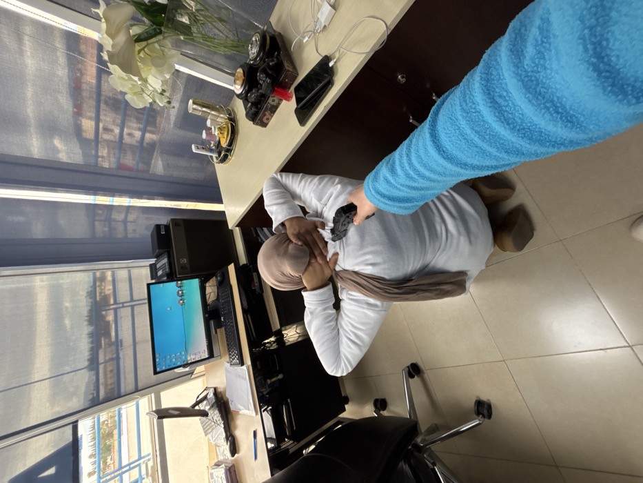The image, captured in Meknes, Morocco, displays an indoor office or workspace environment during daytime. The scene is viewed from an elevated angle, looking down upon two individuals.
In the center of the frame, one person is visible from the back, wearing a white long-sleeved top and a light brown hijab. Their head is slightly tilted forward, and their hands are positioned near their neck or the back of their head. They are situated on a light-colored tiled floor, possibly kneeling or seated. An arm, clad in a bright blue textured long-sleeved garment, extends from the right side of the frame towards this person. The hand holds a black, compact device, which appears to be a massage gun, and is being applied to the upper back or shoulder area of the person in white.
The surrounding setting includes a light-colored desk with visible drawers. On the desk, there is a vase filled with white artificial flowers, a decorative vintage-style camera, a golden circular organizer containing various small items, and a mobile phone connected to white charging cables. To the left of the desk, a computer workstation is set up, featuring a flat-screen monitor displaying a blue desktop background with several icons, a keyboard, and a computer tower. A black office chair with a chrome base is partially visible in the lower foreground. Behind the computer setup, a large window is covered by blue/grey horizontal blinds, allowing a glimpse of outdoor buildings and possibly a street or courtyard bathed in bright daylight.
The image captures an informal moment where one person is providing a back massage or similar therapeutic action to another within what appears to be a standard office setting. No discernible text is visible on any objects or screens.