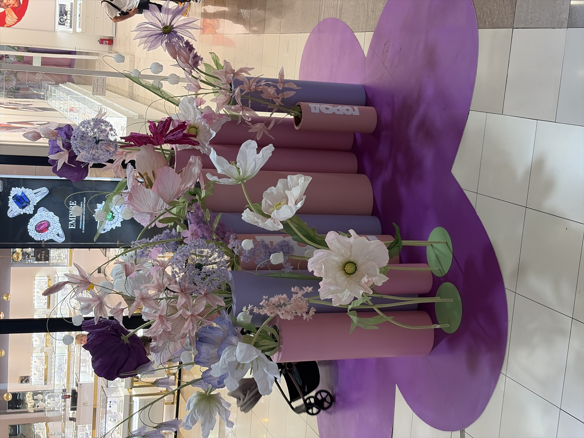 This image depicts a decorative floral display in a commercial setting, possibly a shopping mall. The display features an arrangement of large, artificial flowers in various shades of pink, purple, and white, housed in cylindrical vases of pastel pink and lavender. The flowers are arranged in a tiered manner, with some reaching significant heights. A prominent feature is the purple heart-shaped mat on which the display stands, contrasting with the tiled floor. In the background, through glass storefronts, jewelry displays are visible, suggesting the location is a high-end retail store or a jewelry shop. The overall aesthetic is vibrant and eye-catching. The presence of a baby stroller in the mid-ground hints at a family-friendly environment. A small pink vase with the inscription "город" (gorod, meaning "city" in Russian) is visible on the right side of the display. The lighting suggests an indoor environment, possibly illuminated by overhead mall lighting.