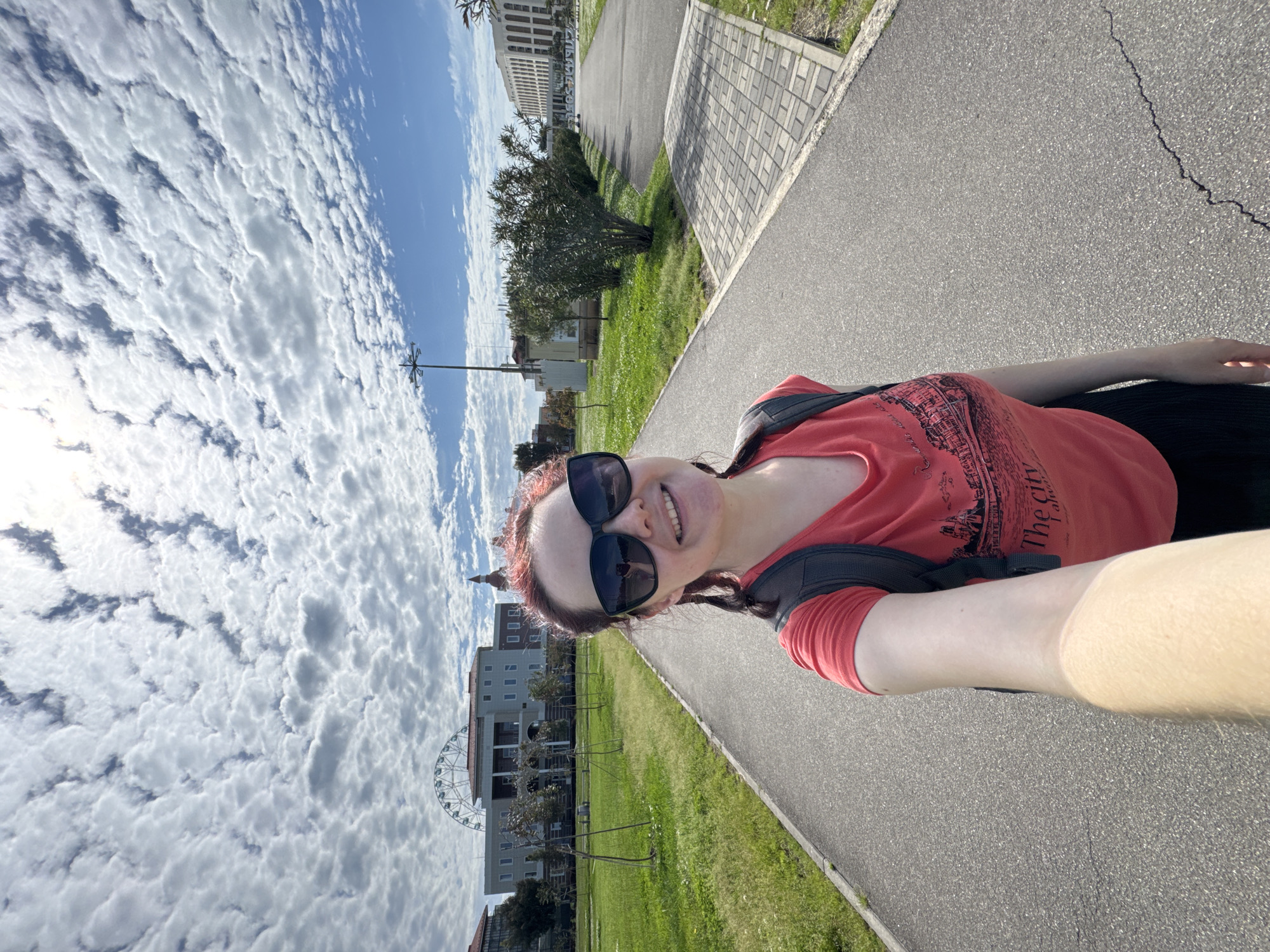 A vertical, medium shot depicts a woman on a paved walkway on a partly sunny day. The woman, wearing a red t-shirt, black sunglasses, and a backpack, smiles at the camera. In the background, a large Ferris wheel is visible, situated in front of a large building. The image suggests the location is a public park or recreational area within the city of Sirius, Russia. The sky is filled with scattered clouds, and the overall scene appears bright. The woman is likely taking a selfie while walking on the paved path.