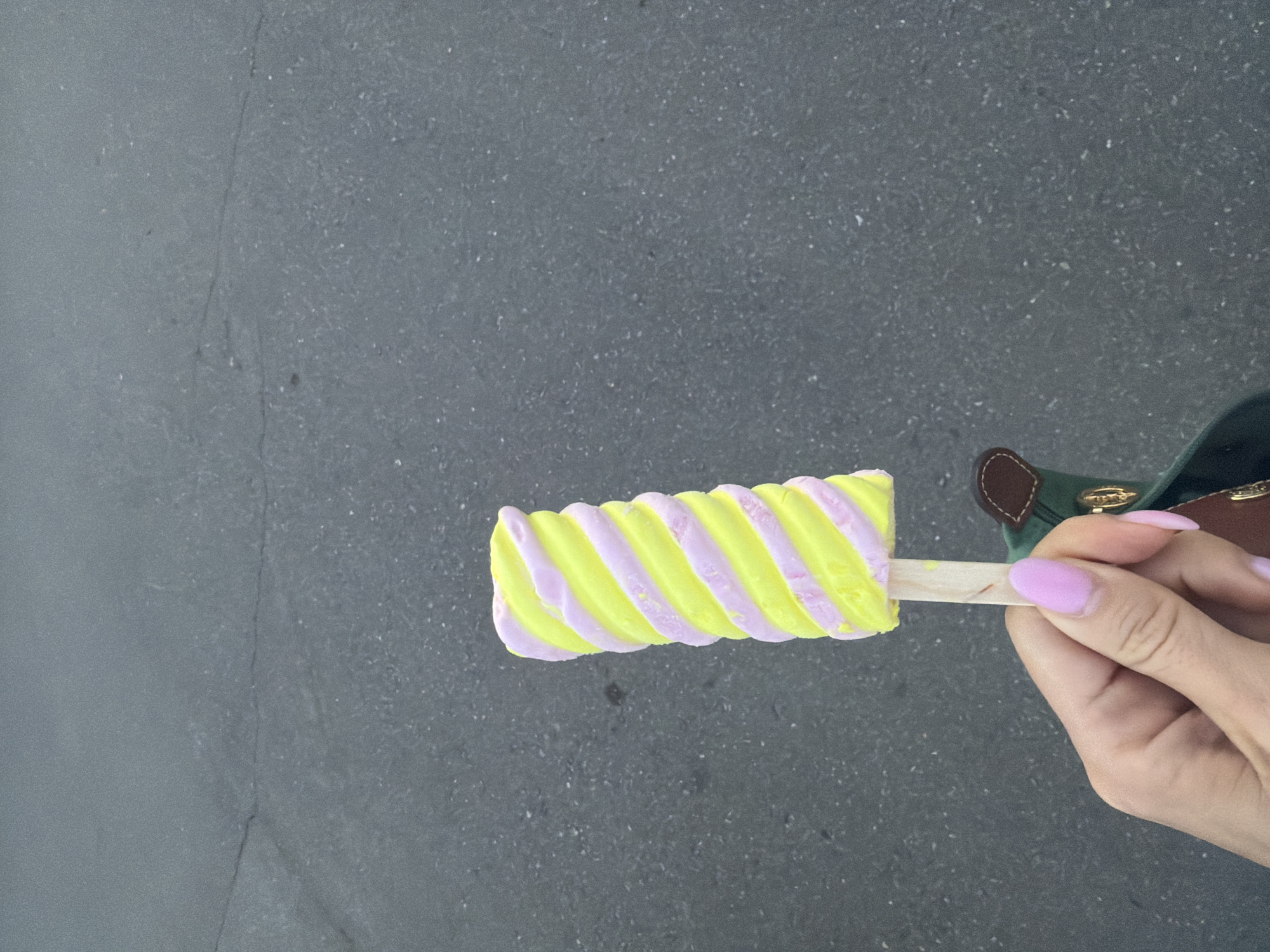 A hand with pink-painted fingernails holds a yellow and pink swirled popsicle on a wooden stick. The popsicle is positioned vertically in the frame, with the top half visible and the bottom half partially obscured by the hand and a dark green bag with brown leather accents. The background is a dark grey, textured asphalt surface, possibly a sidewalk or road, with faint cracks and lighter speckles. There is no visible indication of the location being Moscow, Russia.