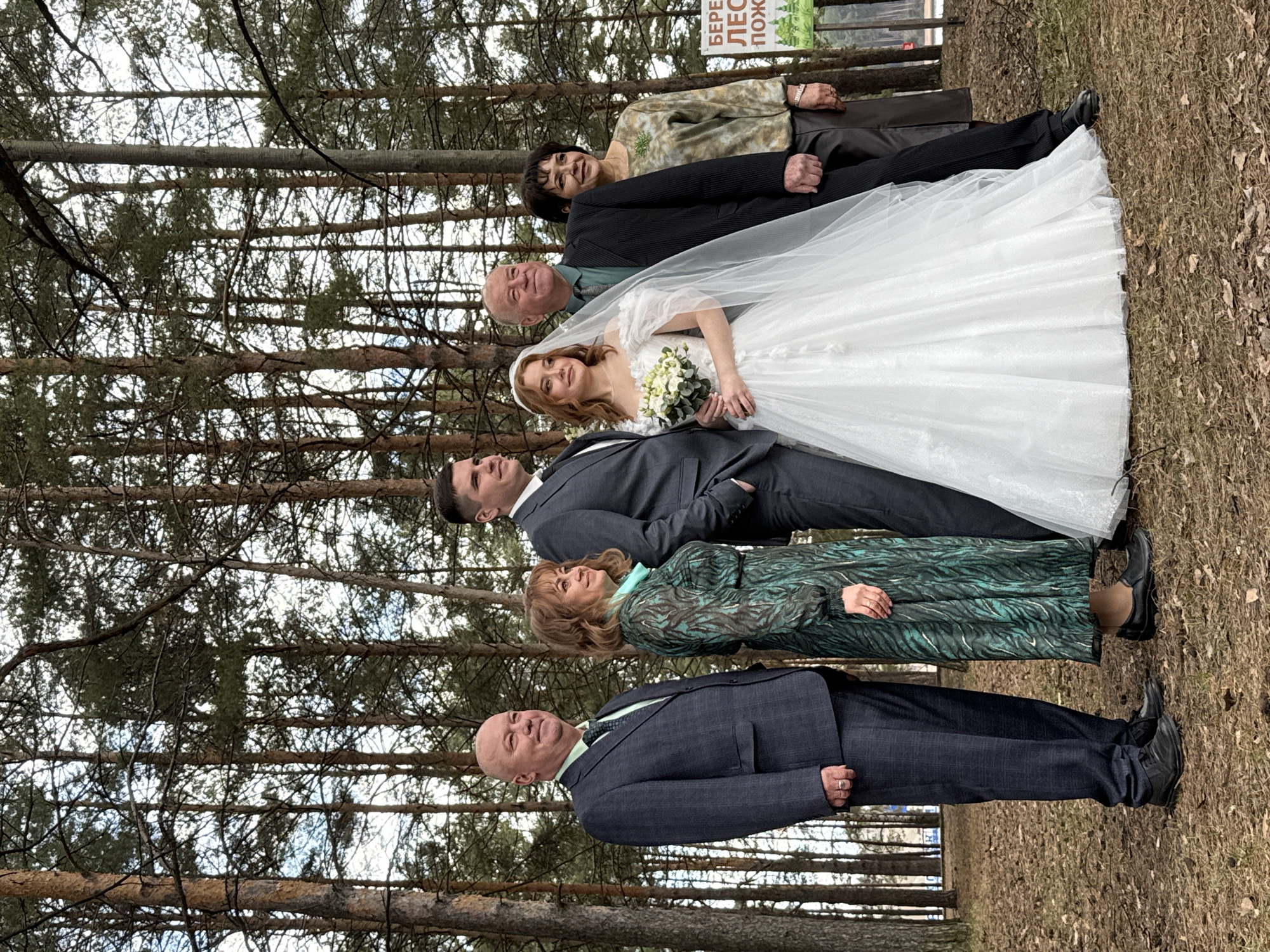 This image captures a group of seven individuals posing for a photograph in a pine forest, likely during a wedding celebration in Chaikovsky, Russia. The group is arranged in a line, with the ground covered in dry leaves and pine needles.
At the center are a bride and groom. The bride wears a white, floor-length wedding dress with a veil and holds a bouquet of white and green flowers. The groom stands next to her in a dark suit. Surrounding them are five other people, presumably family members. On the bride's side, an older man in a dark suit and a woman in a green patterned dress are visible. On the groom's side, an older man in a dark suit, an older woman in a tan and green patterned shirt, and a younger man in a dark suit complete the group. All individuals appear happy, with several smiling.
The setting is a dense pine forest, with numerous tree trunks and branches forming a natural backdrop. The sky, visible through the tree canopy, appears overcast, suggesting daytime with subdued lighting. In the upper right background, a green and white sign is partially visible with Russian text. The legible portion reads "БЕРЕГИ ЛЕС ПОЖА...", which translates to "Protect the forest from fire". The overall atmosphere is celebratory and serene, captured outdoors amidst nature.
