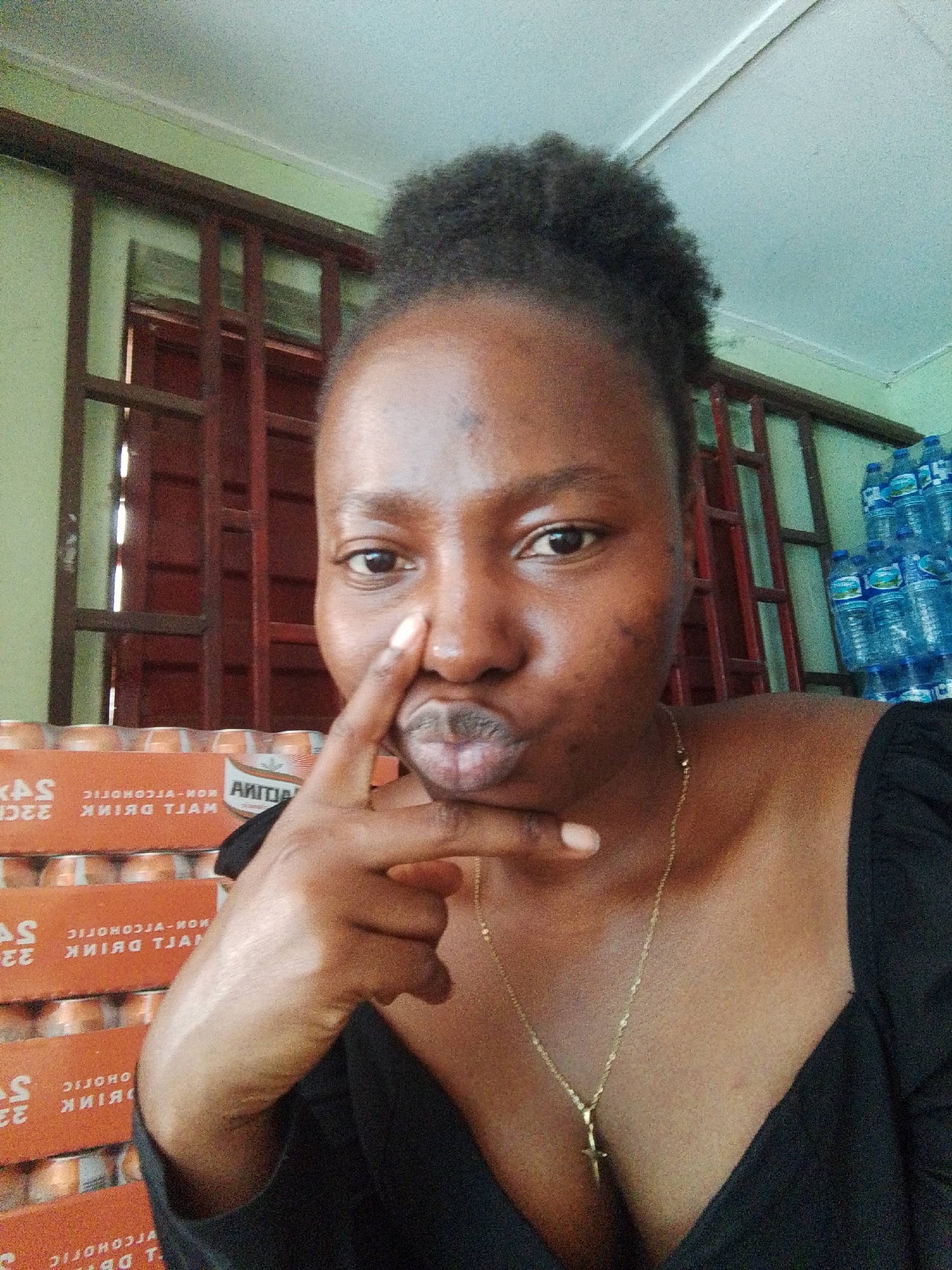 A woman is taking a selfie indoors. She has dark skin, dark hair piled atop her head, and is wearing a black low-cut top and a gold cross necklace. Her index finger is pressed against her upper lip. Stacks of orange cans with the word "KAS" and partially visible text indicating "NON-ALCOHOLIC MALT DRINK" are behind her. To the right are multiple water bottles. A window with a dark maroon frame is behind her, with portions of the wall painted green and light blue. Based on the water bottles and beverage cans visible, the location may be a shop or storage area in Jos, Nigeria.