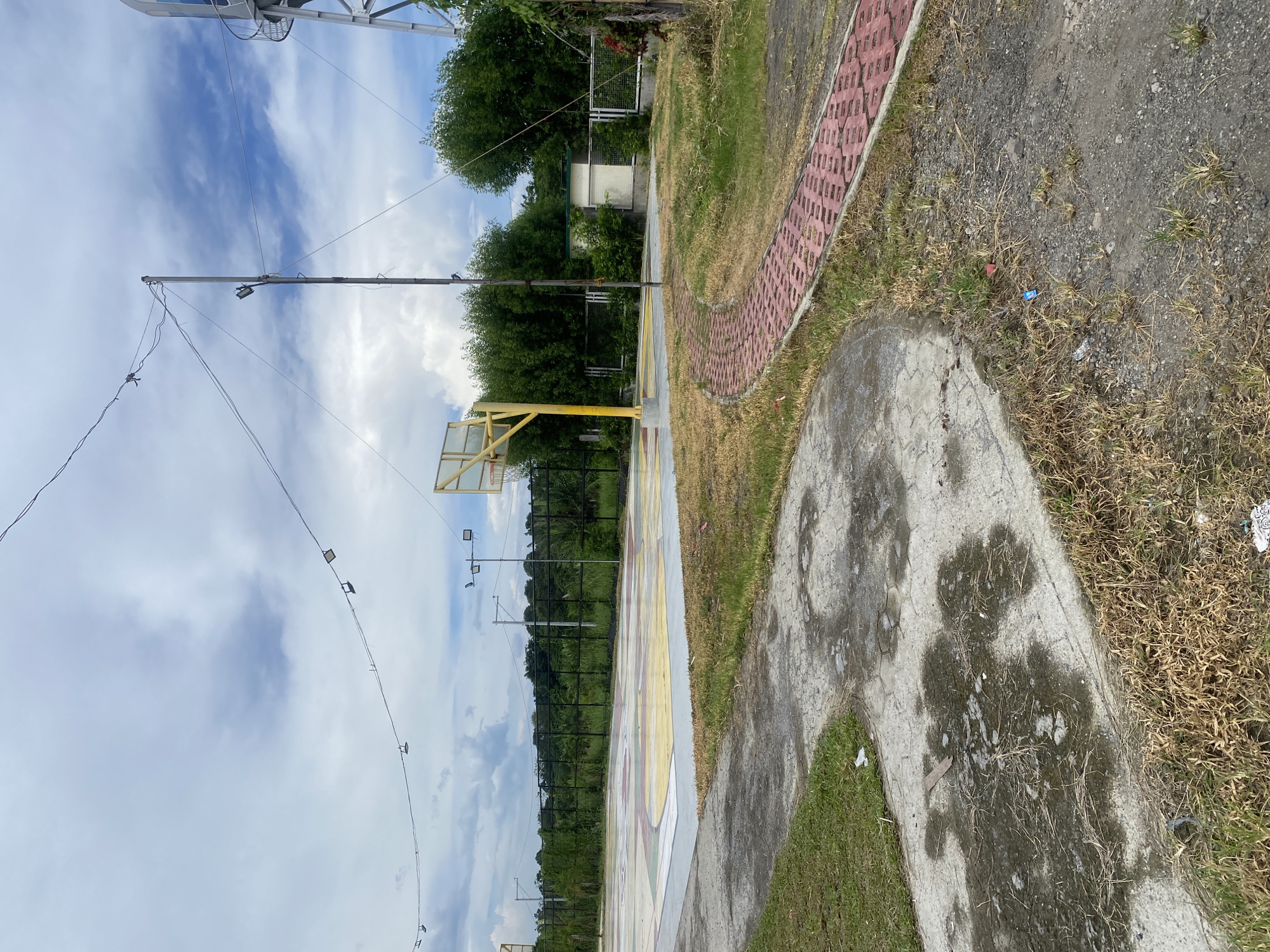 The image depicts an outdoor basketball court located in Davao City, Philippines. The court features a colorful, painted surface with yellow, red, and green sections. A yellow basketball hoop and backboard are visible on the court. A red brick pathway curves around the court, bordered by dry grass and concrete. In the background, there is a black fence, lush green trees, and a water tower. Several utility poles with lights and wires are also present. The sky is overcast with patches of blue. The scene appears to be a public park or recreational area.