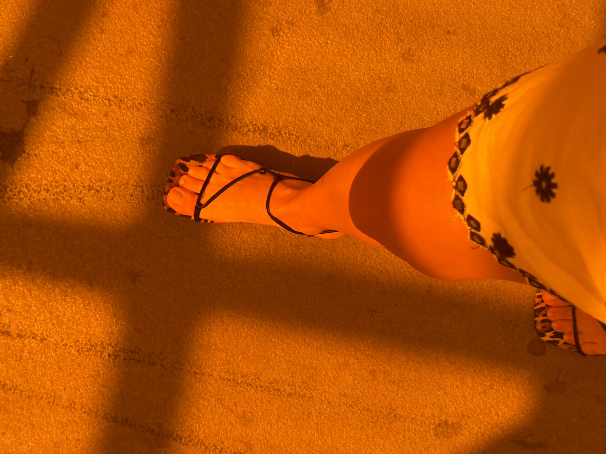 An outdoor, eye-level, full shot captures a person's legs and feet on a textured, sand-colored surface. The person is wearing a yellow dress with a black floral pattern and leopard-print sandals with thin black straps. The scene is dramatically lit by harsh, angled sunlight, casting distinct rectangular shadows across the ground. The overall color palette is dominated by warm oranges and yellows due to the lighting.