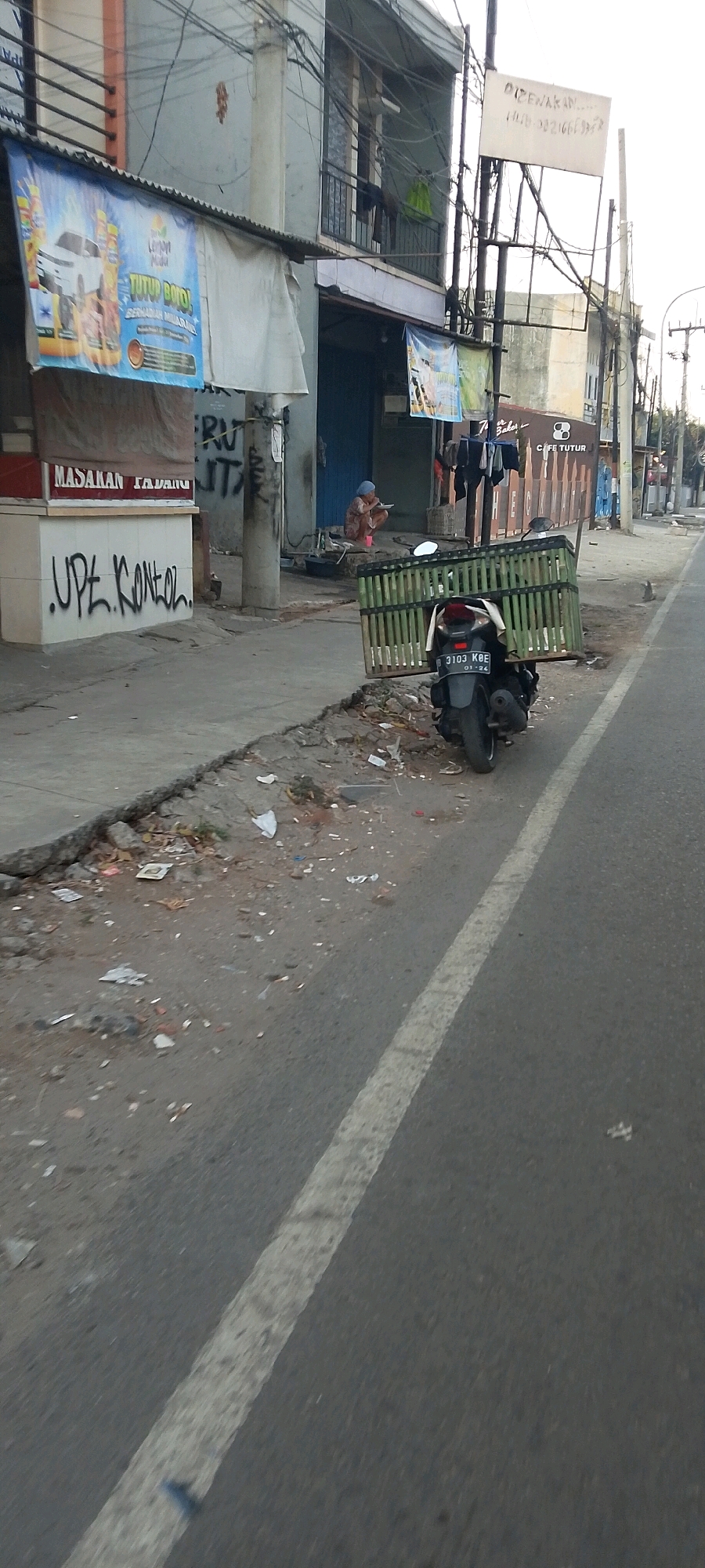 The image depicts a street scene in Bekasi, Indonesia, characterized by a narrow, paved road with a white lane divider. The road is bordered by a concrete sidewalk littered with various debris, including paper, plastic, and other trash.
On the left side of the sidewalk, there is a low building with a metal awning extending over the sidewalk. A banner advertising a product is attached to the awning. Below the awning, a portion of a stall or counter is visible, with text that appears to read "MASAKAN PADANG". Graffiti is present on a concrete pillar and a wall. Further back, a woman wearing a blue headscarf is seated on the ground, appearing to be eating or preparing food.
To the right of the woman, a motorcycle is parked on the sidewalk. The motorcycle is dark-colored and has a large, rectangular bamboo cage attached to the rear. The license plate is partially visible, showing the number "3103 KBE" and "01-24".
Across the street, more buildings are visible, including one with balconies and laundry hanging. A sign with text and a phone number is mounted on a pole. Further down the street, another building has signage that appears to read "CAFE TUTUR". The overall environment suggests an urban setting with a degree of disarray and informal commerce.