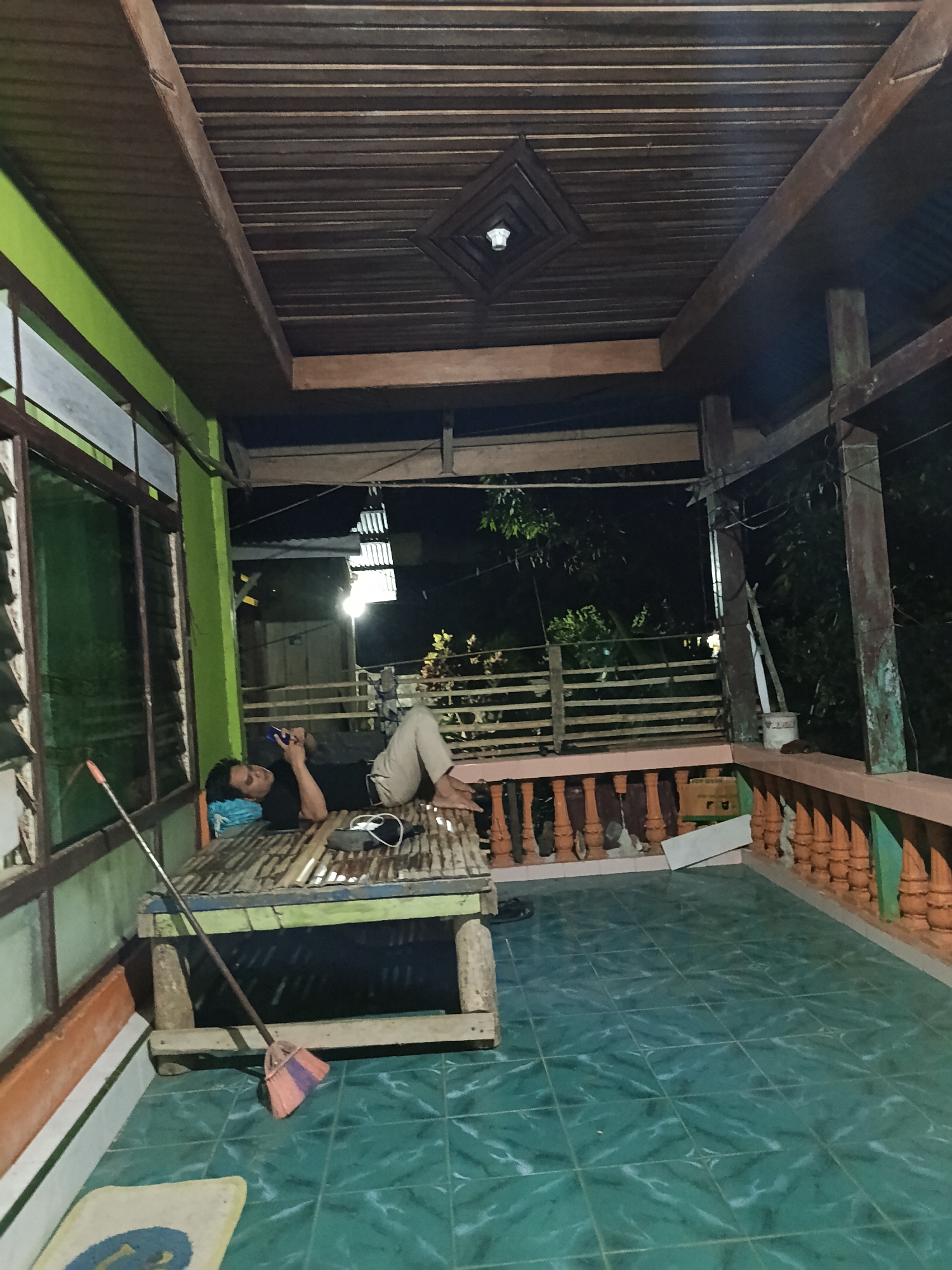 The image shows a person lying on a wooden platform under a covered outdoor area. The person is wearing a black t-shirt and light-colored pants, and is looking at a smartphone. A broom is leaning against the platform. The floor is tiled with a green pattern. The background shows trees and a railing, suggesting an outdoor setting. The architecture suggests it could be in Mansalean, Indonesia.