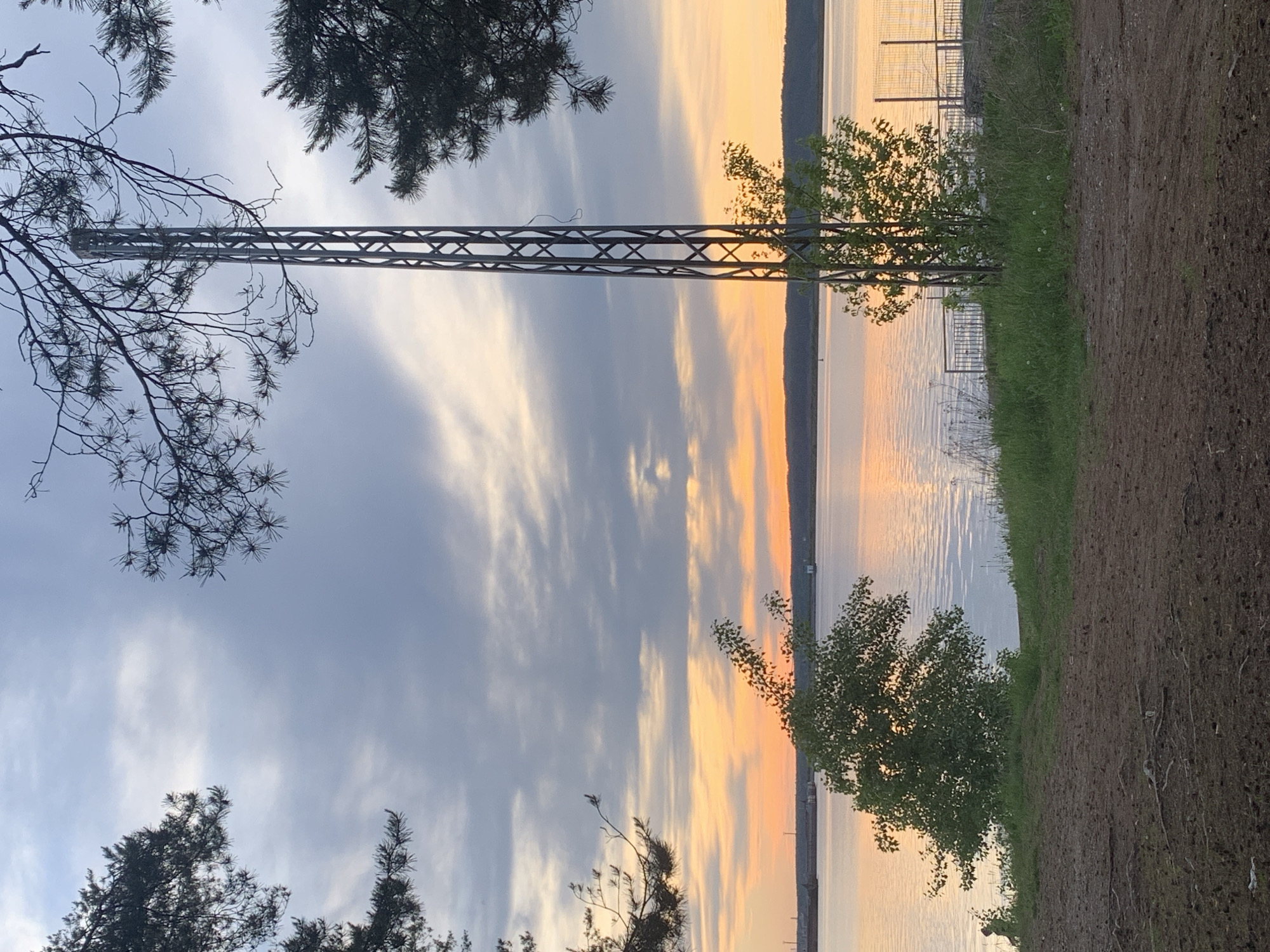 The image depicts a landscape at sunset or sunrise, likely near a body of water. The composition is framed by trees, with a metal structure running vertically through the scene. To the right, there is grass and dirt, suggesting a shoreline or embankment. In the distance, a fence-like structure is visible along the water's edge. The sky displays a mix of blues, greys, and orange hues, indicating the time of day. Based on the mention of Chaikovsky, Russia, the location is likely near a lake or river in the area, such as the Kama River.