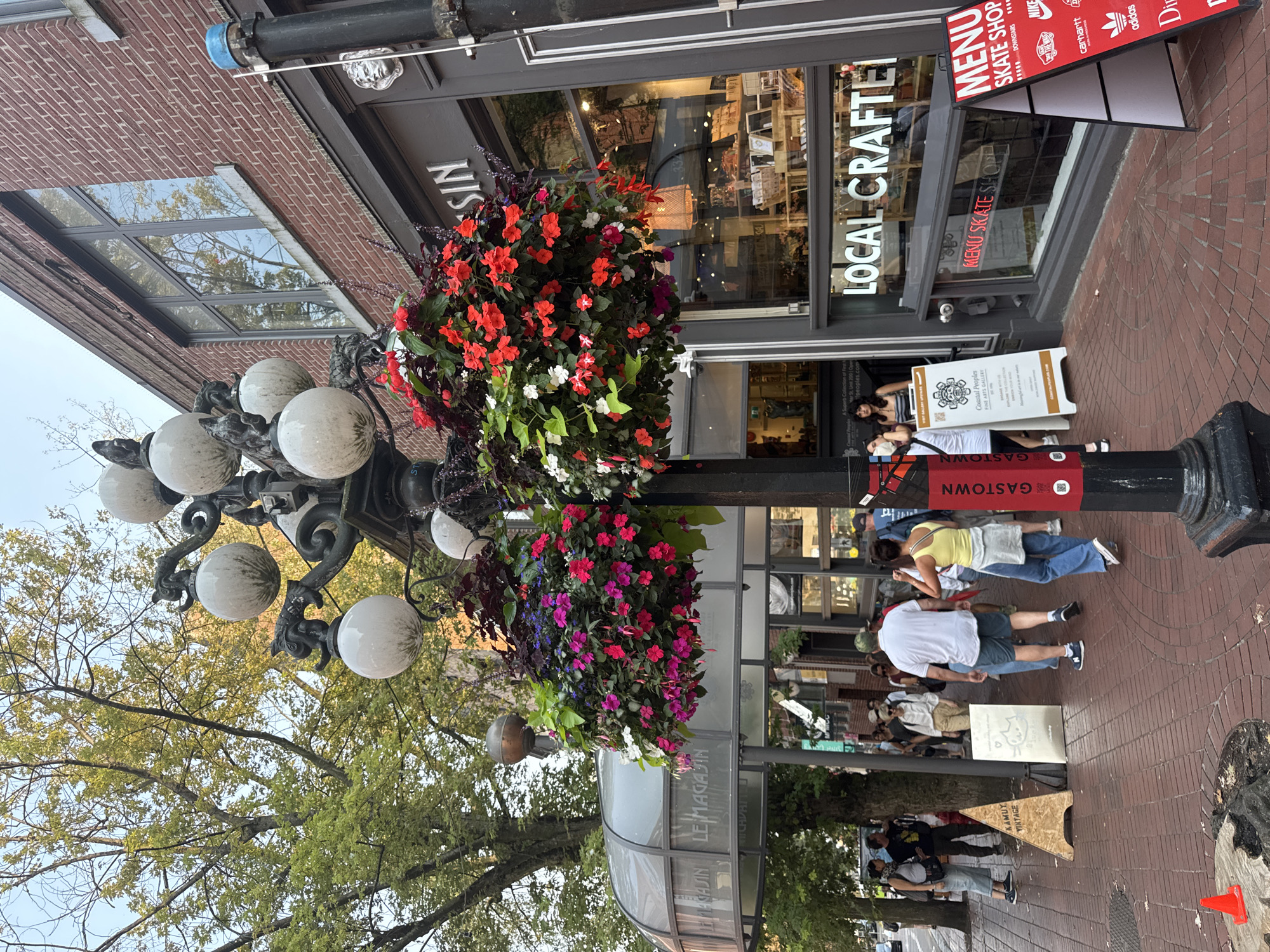 This image depicts a street scene in Gastown, Vancouver, Canada. A lamppost with multiple globe lights and hanging flower baskets is prominently featured in the foreground. The baskets are filled with vibrant red, pink, and purple flowers, along with lush green foliage. To the right of the lamppost is a building with a brick facade and large windows. A sign above one of the windows reads "Local Crafted". In front of the store, an A-frame sign displays "MENU SKATE SHOP" with logos of various brands like Nike, Adidas, and Carhartt.
On the left side of the image, there is a covered walkway or entrance with "LE MAGASIN" text visible on the glass. A group of people are walking on the brick sidewalk. Some are tourists, identifiable by their casual attire and backpacks. A wooden A-frame sign with "KAMUY VINTAGE" is placed on the sidewalk. The scene is set during the daytime, with natural light illuminating the area. The overall atmosphere suggests a bustling urban environment with a mix of shops and pedestrian activity.