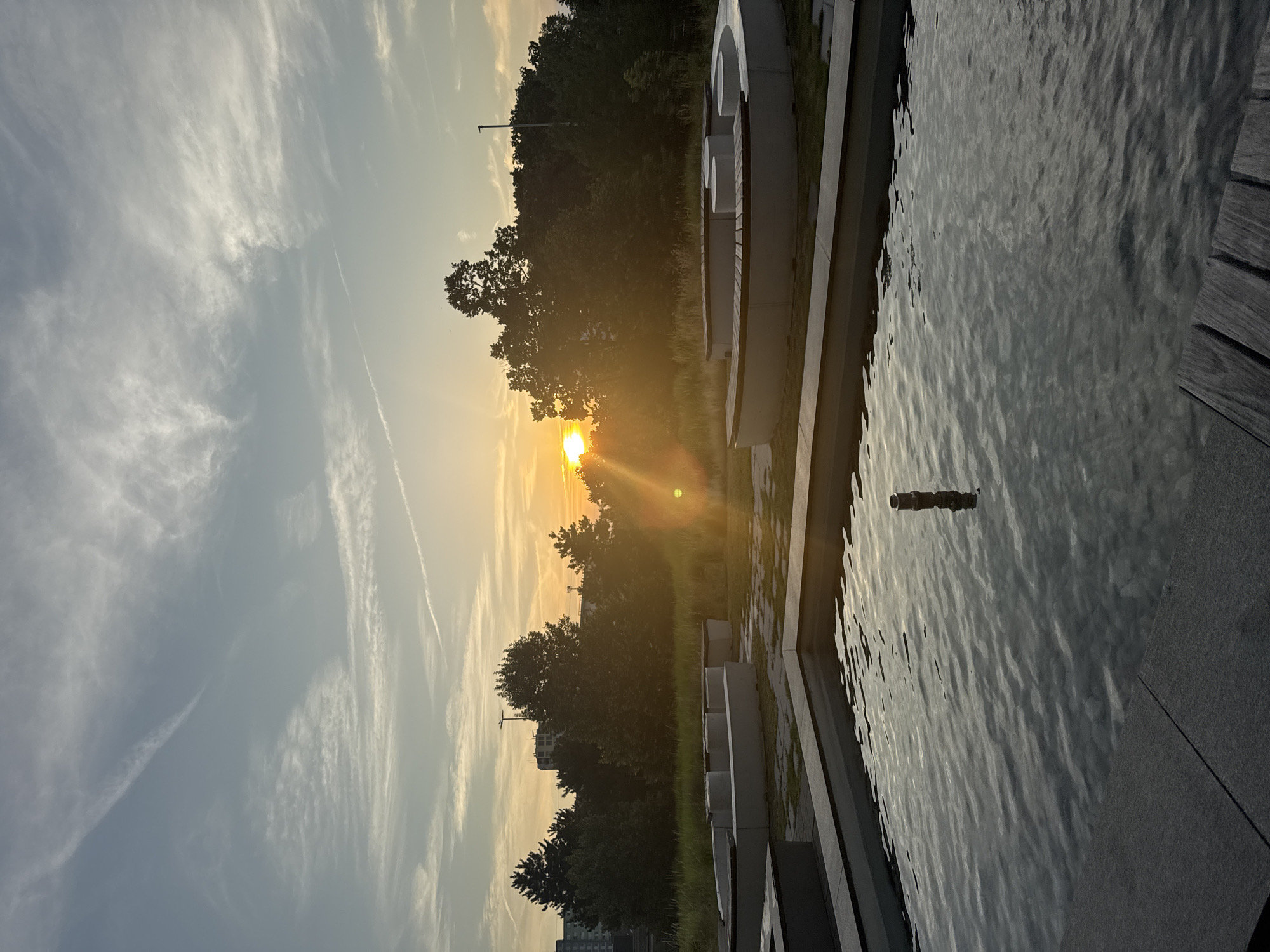 This image captures a sunset scene in Bratislava, Slovakia, likely at the Eurovea waterfront. In the foreground, a modern water feature with dark stone edging and a shallow pool reflects the sky and surrounding elements. The water is gently rippling, revealing submerged pebbles and a dark, cylindrical object emerging from its surface. Beyond the pool, a seating area with curved, white concrete benches and wooden slats is visible, set against a backdrop of lush green grass and mature trees. The trees form a dense line, silhouetted against the vibrant sunset. The sun, a bright orange orb, is low on the horizon, casting a warm glow and lens flare across the scene. The sky above is a blend of pale blue and soft orange hues, with wispy white clouds and contrails adding texture. In the distance, the outlines of city buildings are faintly discernible behind the trees, suggesting an urban environment adjacent to this landscaped outdoor space. The overall atmosphere is serene and tranquil, highlighting the interplay of natural light and contemporary design.