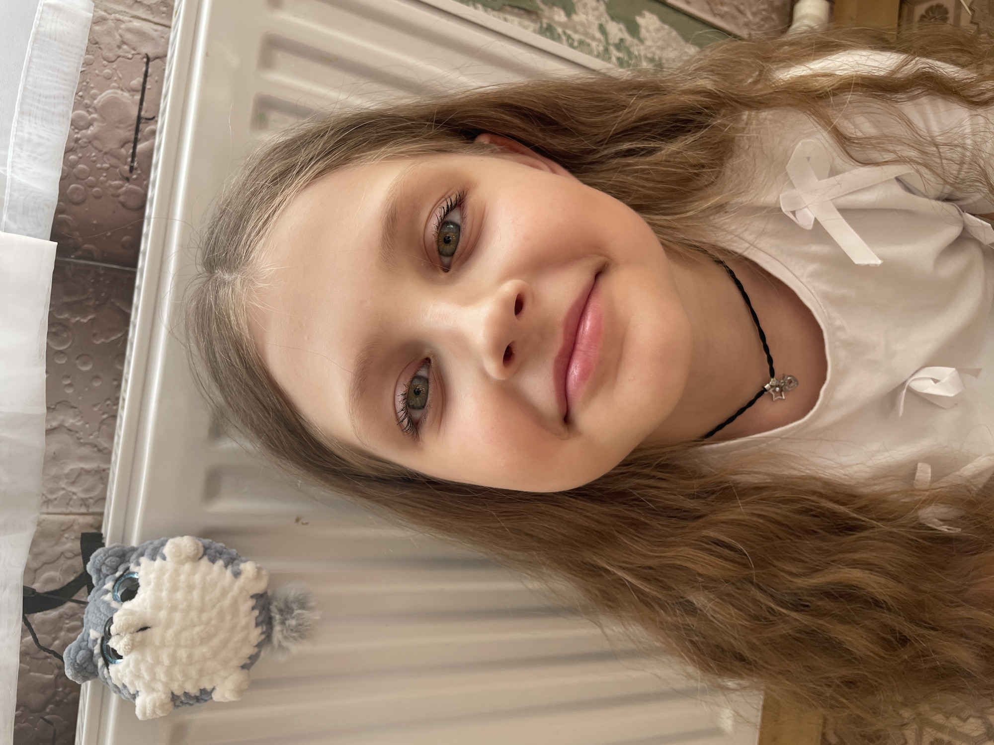A young girl with light brown hair, green eyes, and a small smile is the primary subject of this photograph. She is wearing a white shirt with ribbon embellishments and a thin black necklace with a small, light-colored charm. A crocheted stuffed animal resembling a gray and white cat lies to her left. The background consists of a white radiator, part of a curtain, and a textured wall. Location details in Izhevsk, Russia, are not discernible.