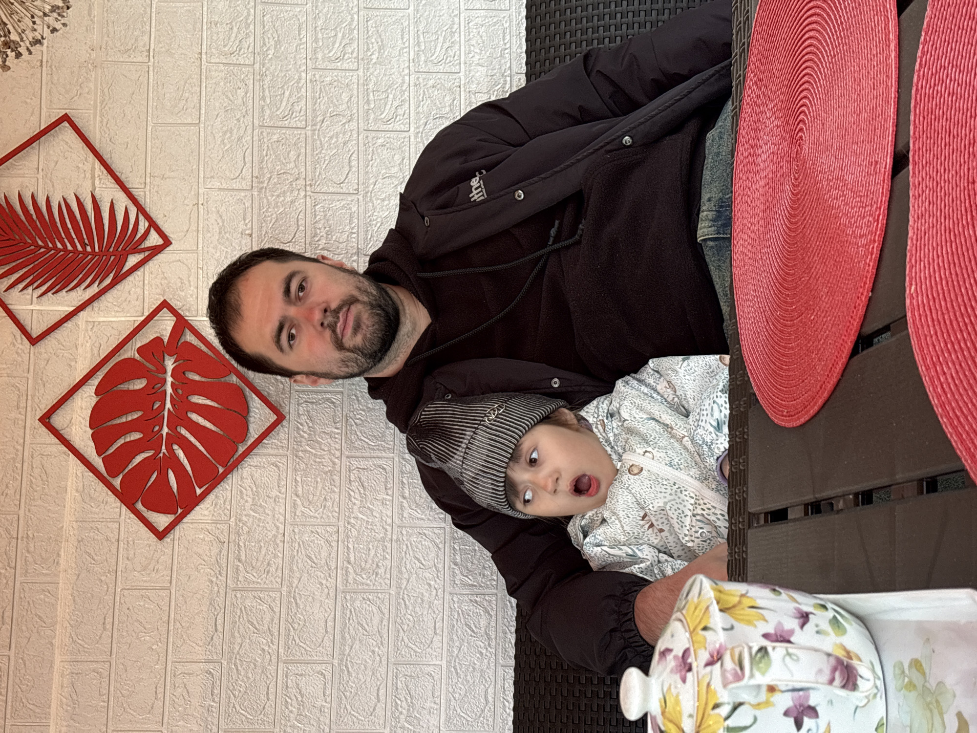 The image captures a man holding a child on his lap at an outdoor table. The man has dark hair and a beard, wearing a black jacket and hoodie. The child wears a striped hat and patterned jacket with an open mouth. They are seated at a table with red woven placemats and a floral teapot. The background features a textured white wall with two red framed leaf decorations. This image was most likely taken in a private residence in the city of Chaikovsky, Russia.