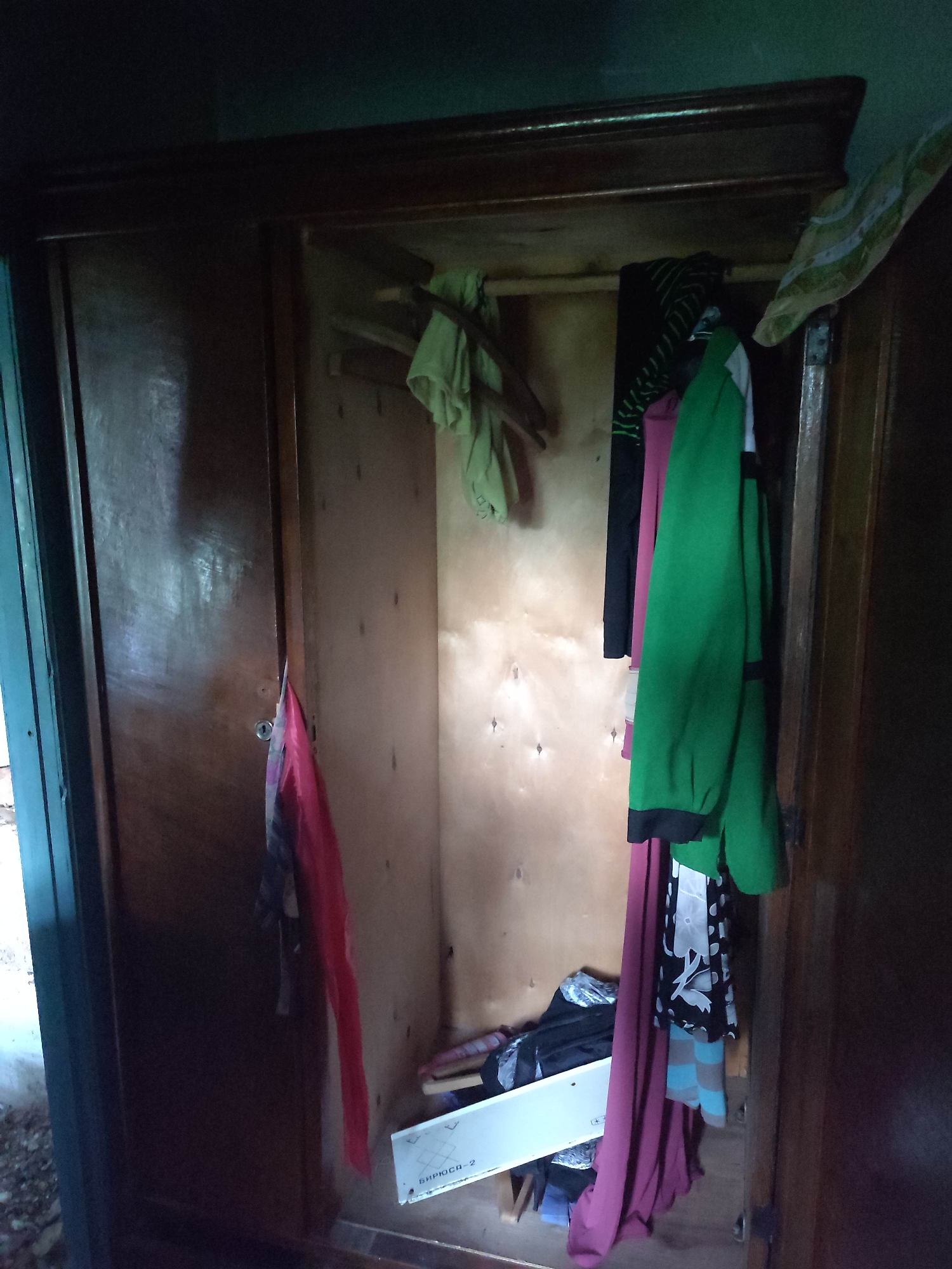 The image shows the interior of a wooden wardrobe, likely located in Gremyachinsk, Russia, due to the context provided. The wardrobe is filled with various items of clothing hanging from a rail and piled on a shelf. On the left side, a pink garment is draped over the wardrobe door. To the right, several articles of clothing are hanging, including a green jacket with black trim, a black and white patterned top, and a pink dress or skirt. A wooden shelf inside the wardrobe holds a white box with Cyrillic writing and some folded clothing. The overall environment appears dim and somewhat cluttered.