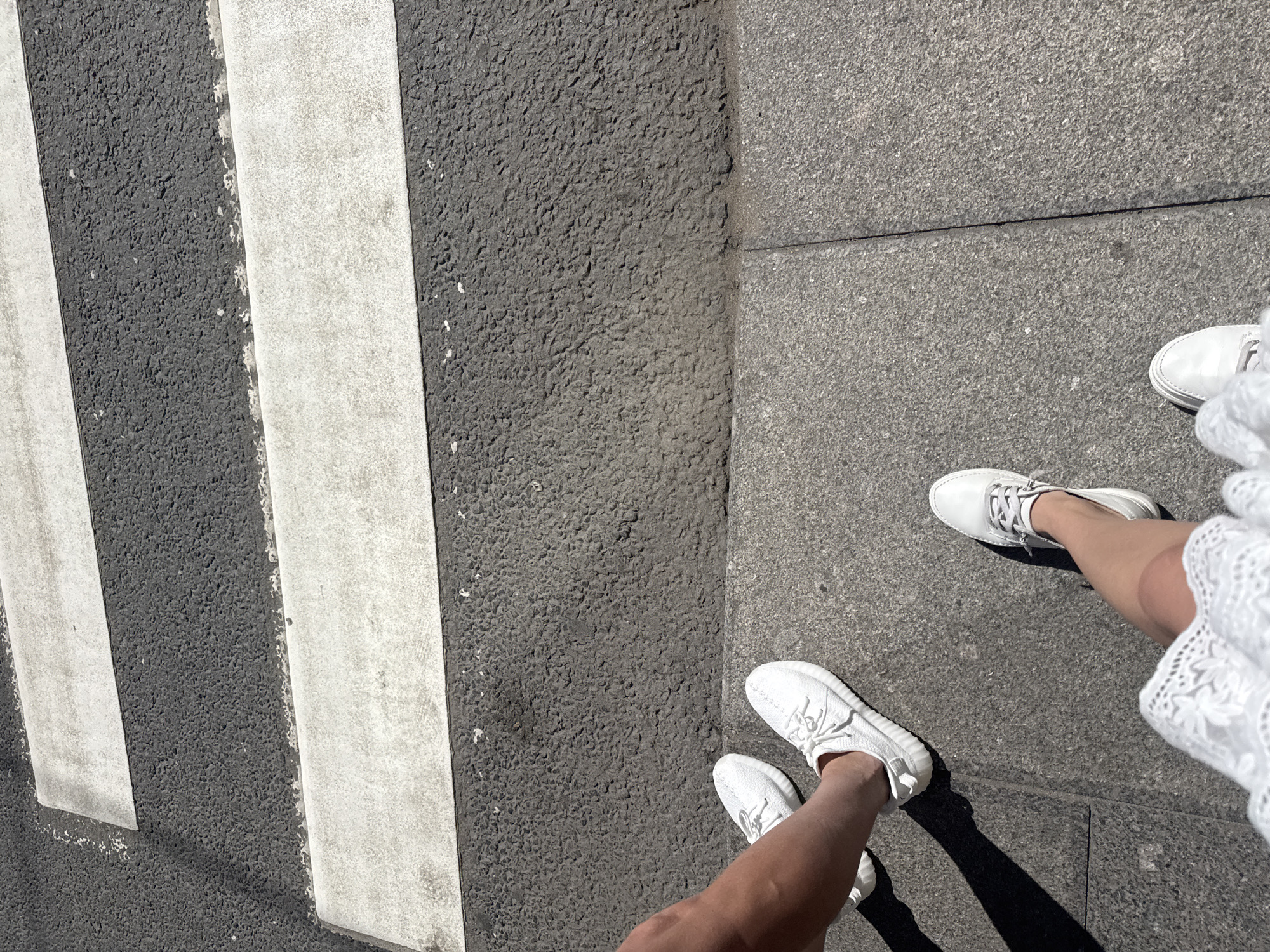 This is a ground-level, close-up, eye-level shot of two people standing on a sidewalk next to a crosswalk. The crosswalk features the characteristic white stripes seen on streets in Saint Petersburg, Russia. The surface of the crosswalk is asphalt, with visible white paint for the pedestrian markings. The sidewalk is made of large, grey stone slabs. One person, on the left, is wearing white sneakers and has their bare leg exposed, suggesting warm weather. The second person, on the right, is wearing white shoes and a white lace dress. Both individuals are facing away from the camera, looking towards the crosswalk. The lighting suggests a bright, sunny day, casting distinct shadows. The scene captures a moment of pause before crossing the street.