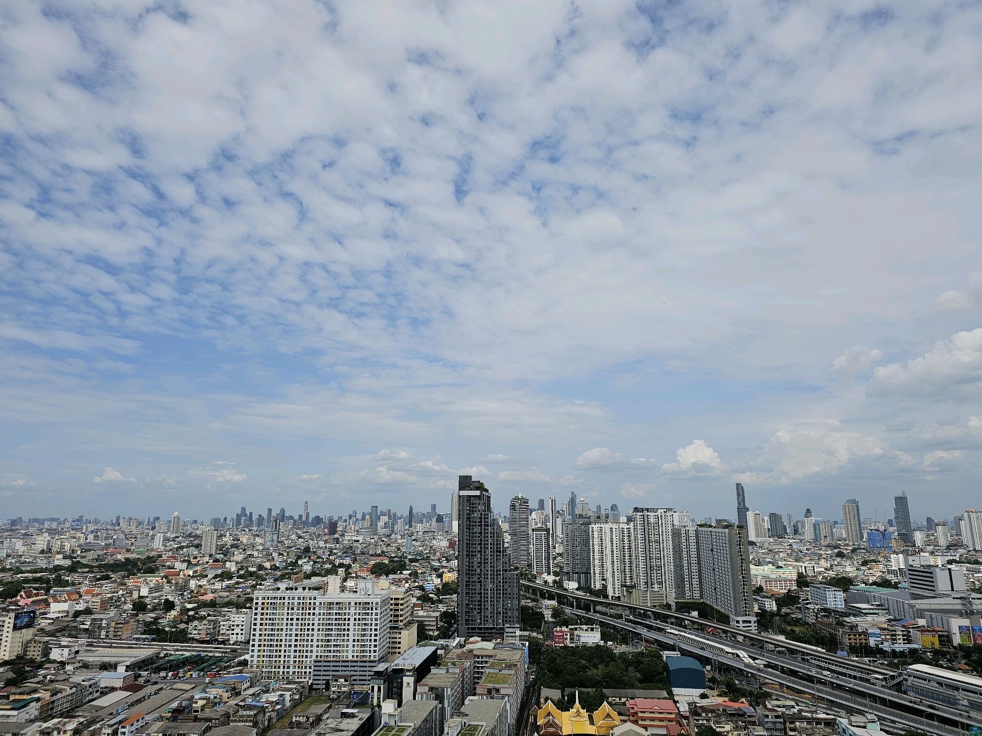 This is a high-angle, wide-shot photograph of a sprawling cityscape, likely taken from a tall building. The scene is dominated by a vast urban landscape with numerous buildings of varying heights and architectural styles. In the foreground, several mid-rise residential or commercial buildings are visible, some with prominent balconies. A large, white, modern apartment building with a grid-like facade stands out to the right. Further into the city, a dense collection of buildings stretches towards the horizon, with a cluster of taller skyscrapers forming a distinct skyline in the distance. Elevated train tracks and roadways weave through the city, indicating a well-developed transportation network. The sky above is a pale blue, partially covered by a textured layer of white and grey clouds that are somewhat scattered, creating a dynamic and slightly overcast atmosphere. The overall impression is of a densely populated, modern metropolis with a mix of older and newer construction. The location is identified as Bangkok, Thailand.