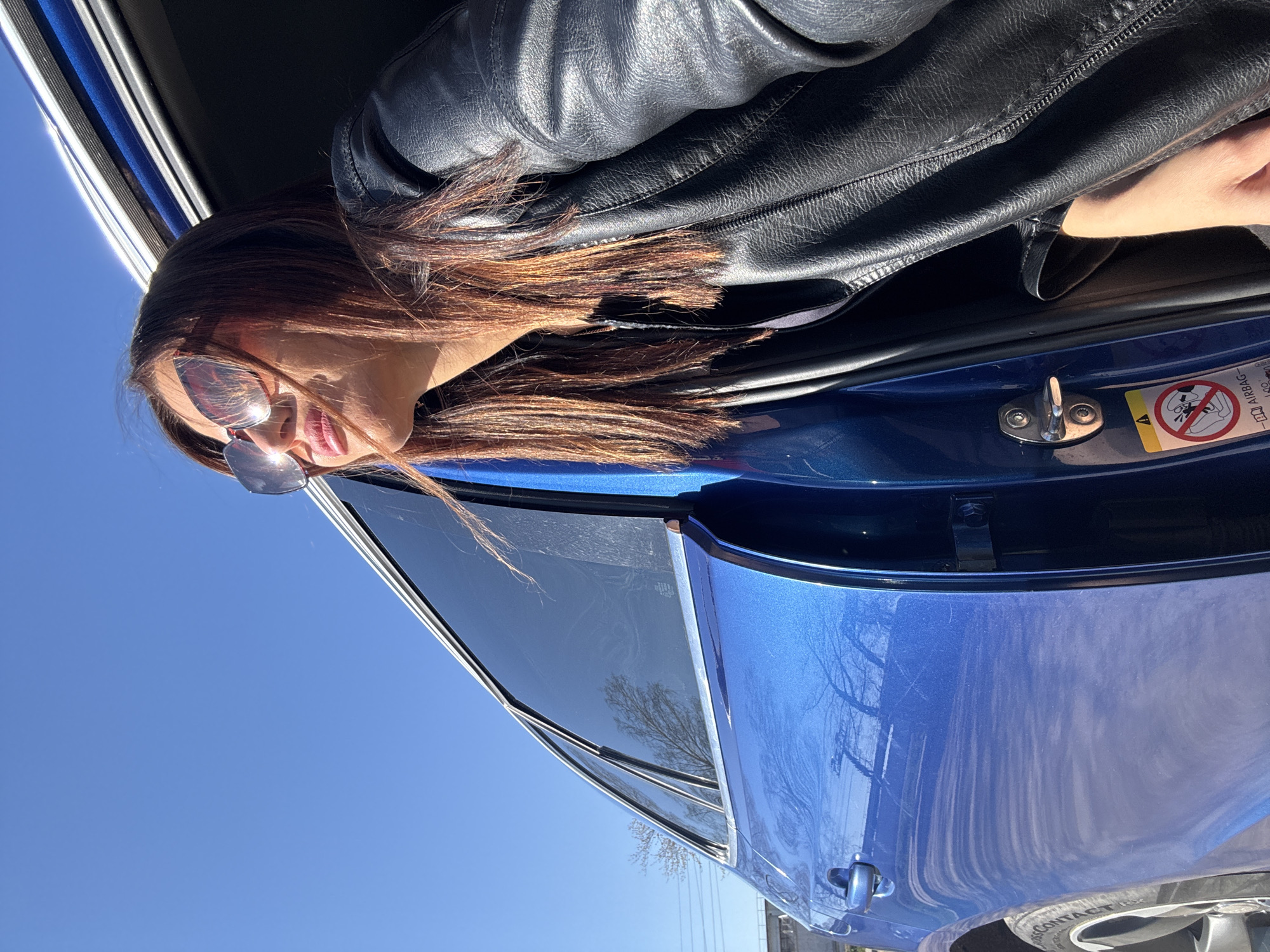 A woman wearing a black leather jacket and sunglasses leans out of the open door of a blue car under a clear, bright blue sky. The location is unknown and there is no evidence to suggest it is in Chaikovsky, Russia.