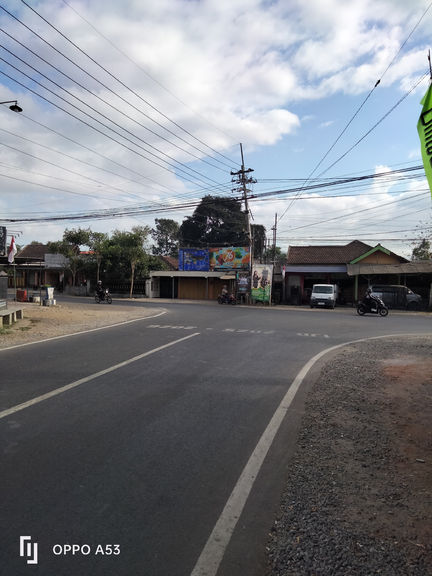 The image depicts a street scene in Wonoanti, Indonesia, under a partly cloudy sky. A paved road curves from the foreground into the midground, with a white line marking its edge and a dashed white line indicating traffic lane separation. On the left side of the road, a gravel shoulder leads to a sidewalk with a stone bench. Beyond the sidewalk are buildings with tiled roofs and storefronts, some with signage and posters. One poster features an Indonesian flag. A motorcycle is parked on the left side of the road.

On the right side of the road, the gravel shoulder continues and transitions to a dirt area with scattered rocks. Further back, there are more buildings with tiled roofs and some vegetation. A white van is parked in front of one building, and a black motorcycle is visible in front of another. Several utility poles with numerous overhead wires crisscross the sky above the street. A green flag with white text is prominently displayed on the right side of the image, attached to a pole. A streetlamp is visible on a pole on the far left. The overall impression is a functional, everyday street in a semi-urban or rural Indonesian setting.