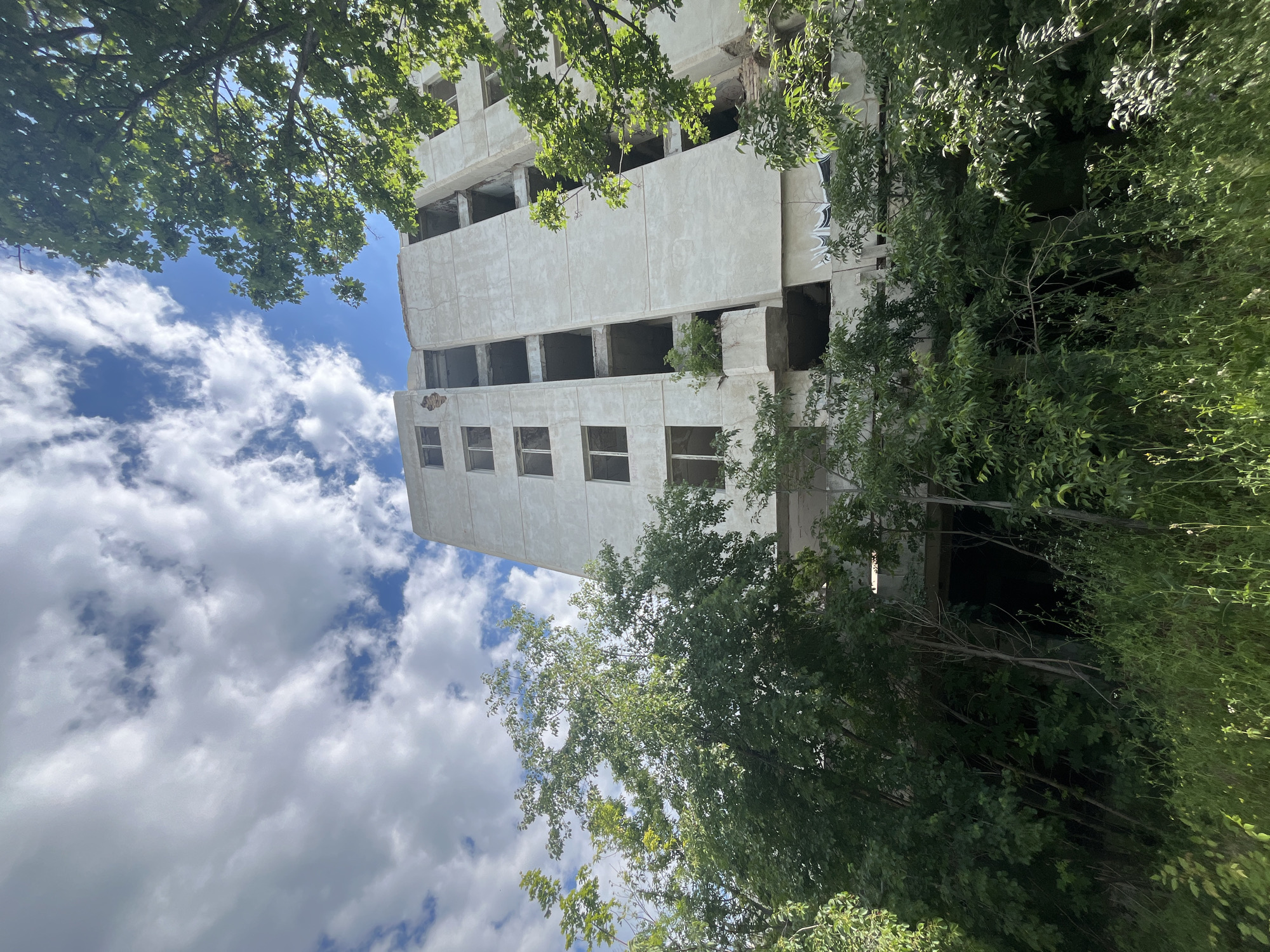 A low-angle shot captures an abandoned, concrete apartment building overgrown with lush green trees and foliage, set against a backdrop of a bright blue sky dotted with fluffy white clouds. The building, appearing weathered and possibly structurally compromised with visible damage and missing windows, is partially obscured by the dense vegetation. The scene evokes a sense of decay and nature reclaiming an urban structure, likely located in Varna, Bulgaria, a city known for its coastal landscape and historical architecture. There are no people or animals visible in the image.