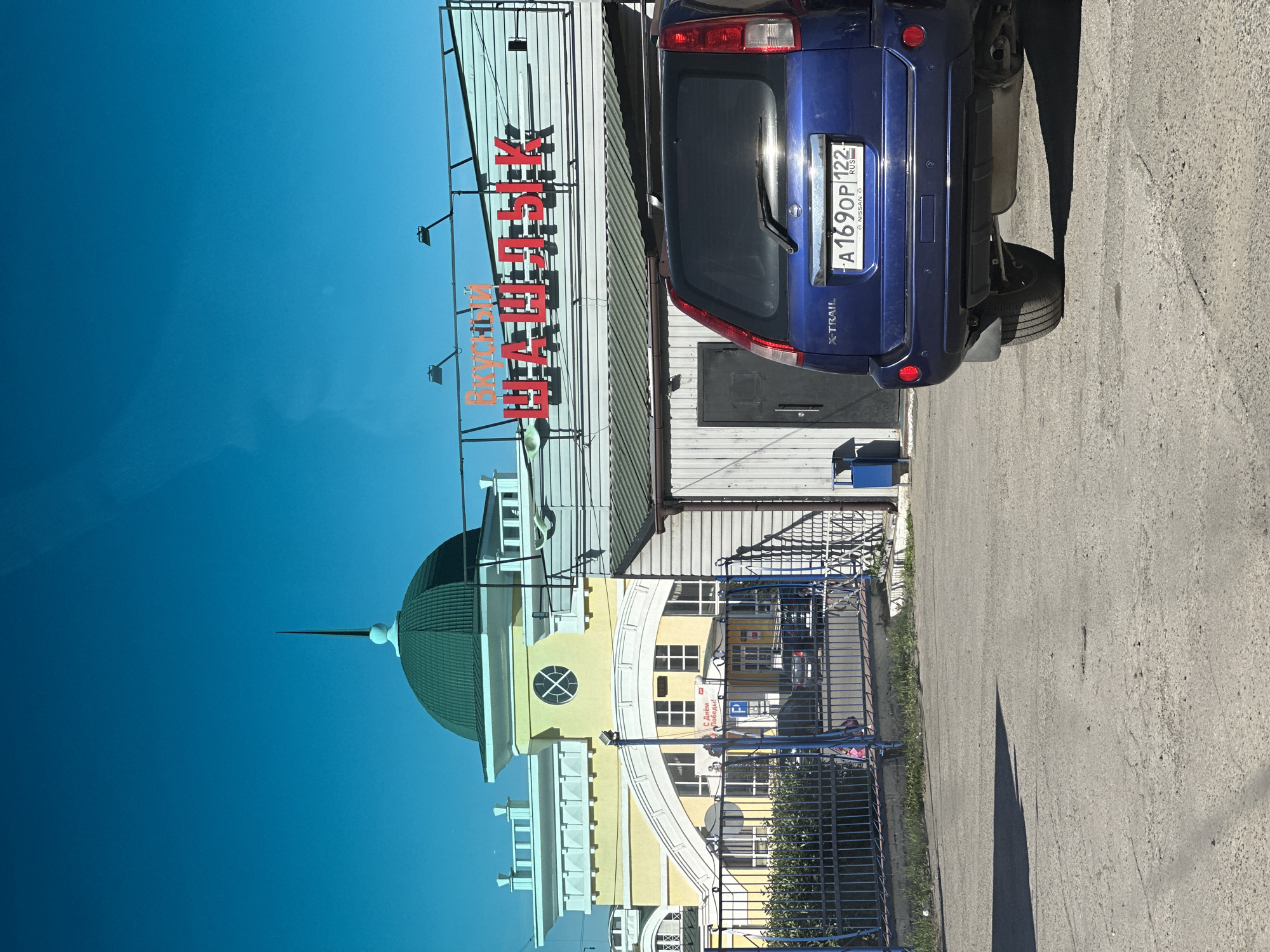 The image shows a blue Nissan X-TRAIL parked in front of a building with a green dome. The building has a sign that reads "Вкусный Шашлык" (Tasty Shashlik). The license plate of the car is visible and reads "A 169OP 122". In the background, there is a metal fence with a sign that says "С Днем Победы!" (Happy Victory Day!). The scene appears to be in a public area with some vegetation and a paved surface. The sky is a clear blue. The context suggests this is likely a cafe or restaurant in Novoaltaysk, Russia, specializing in shashlik, a type of grilled meat.