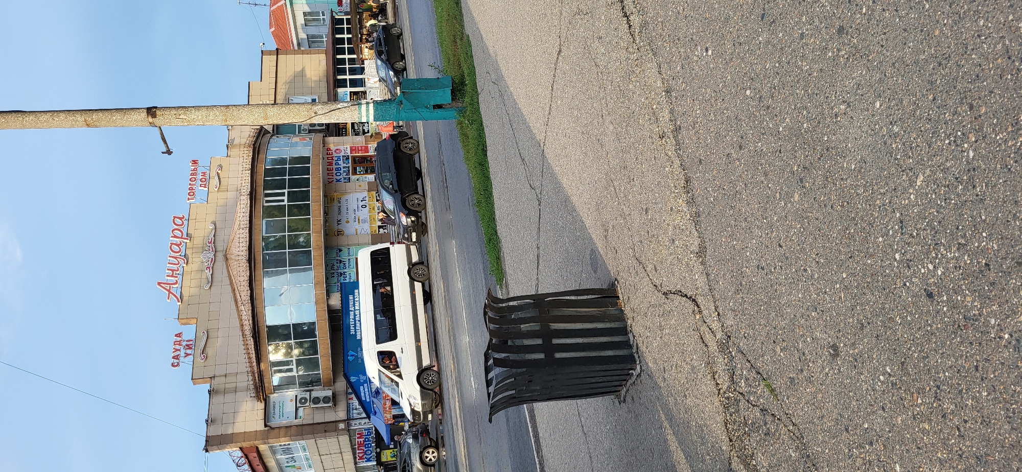 The image shows a street scene in Oskemen, Kazakhstan, with a commercial building named "АНУАРА" and "ТОРГОВЫЙ ДОМ" on its facade. Several vehicles, including a white minivan and black SUVs, are driving on the road. A black metal trash receptacle is positioned on the sidewalk in the foreground, and the pavement shows signs of cracking. The sky is blue with some clouds, and sunlight casts shadows on the ground. The scene appears to be during the daytime.