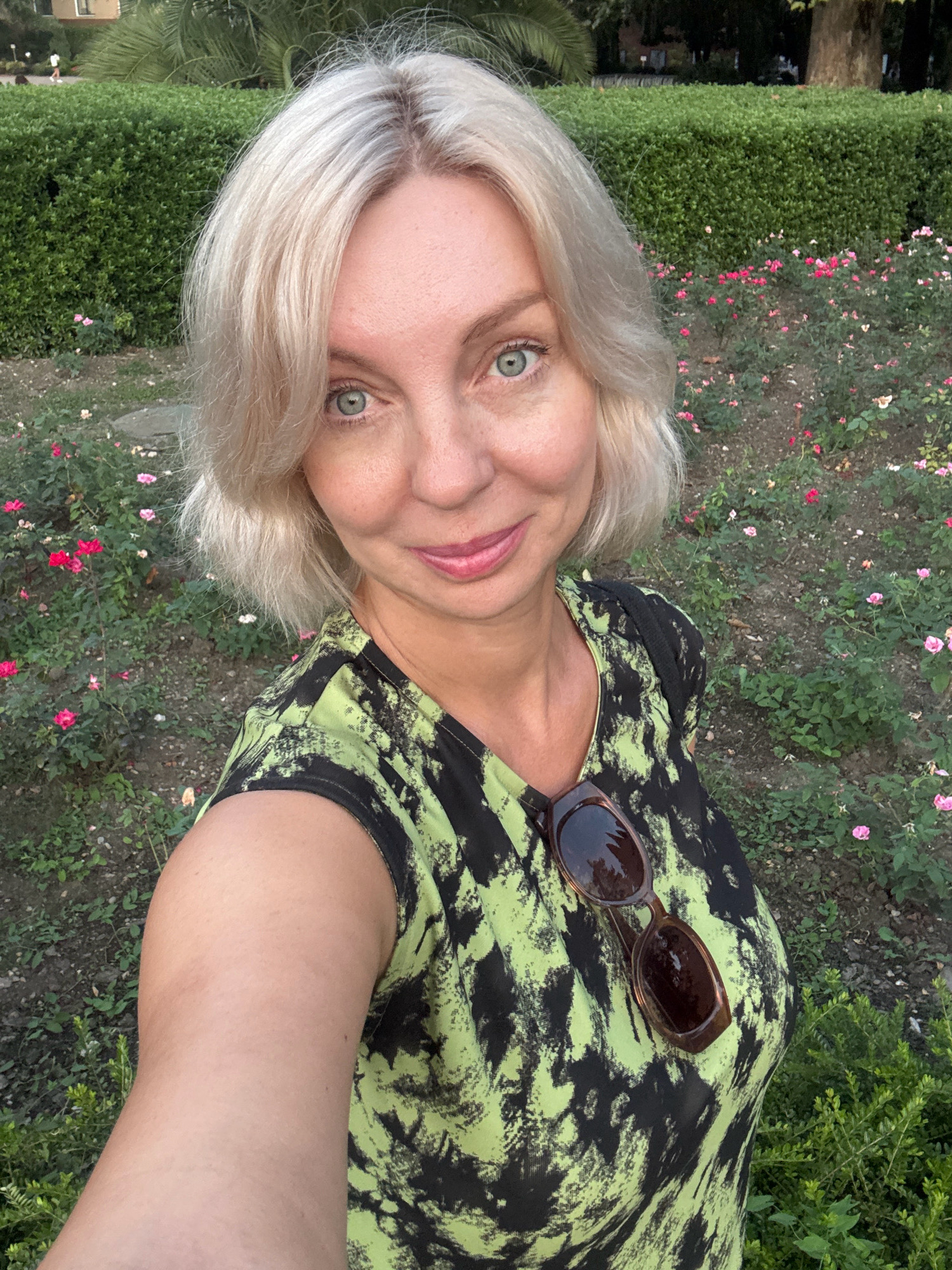 A woman with blonde hair, wearing a green and black patterned shirt and sunglasses hanging on her shirt, smiles at the camera. She is outdoors in a garden with rose bushes and a trimmed hedge. This scene is located in Sochi, Russia, where the climate is subtropical.